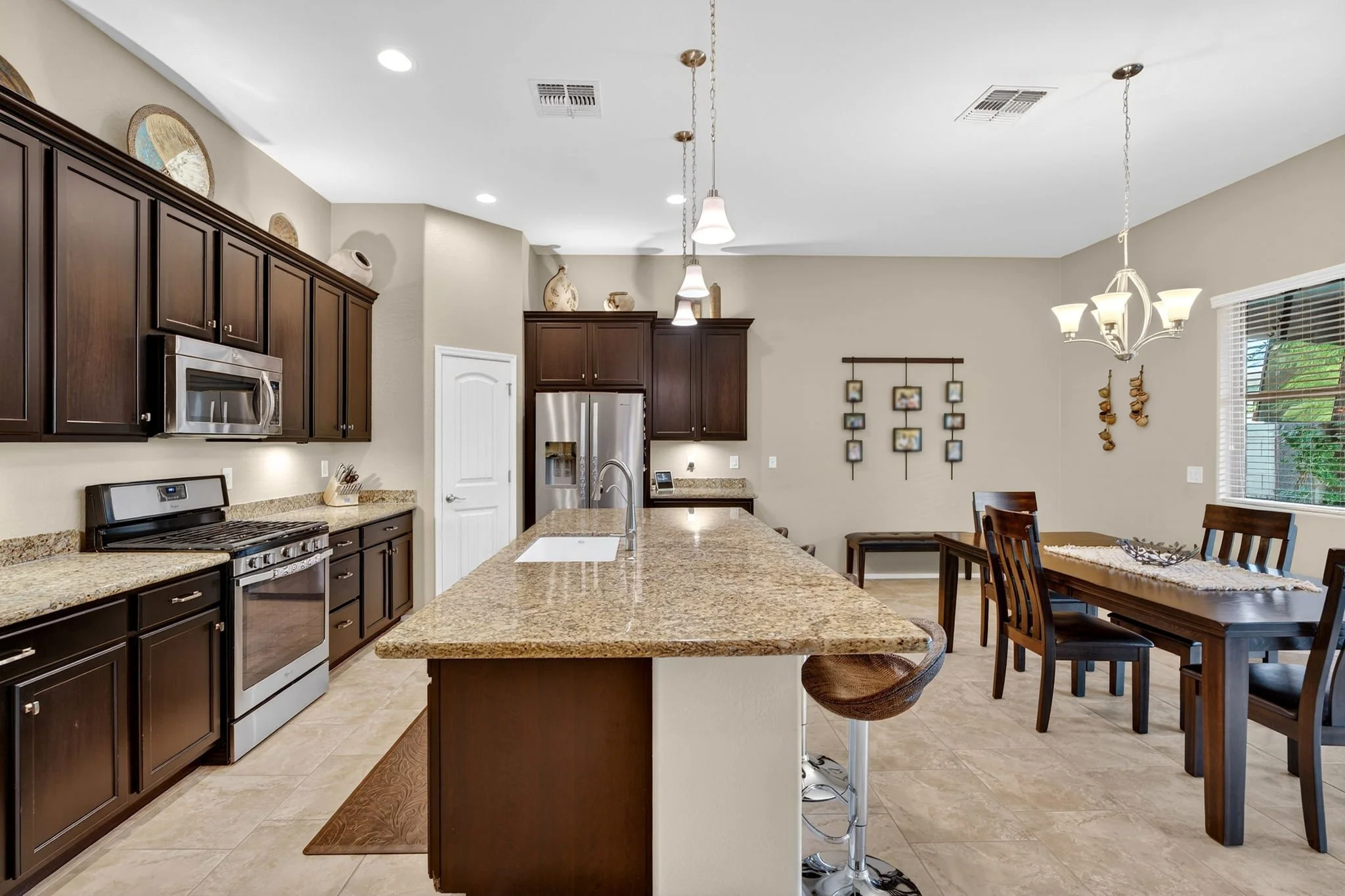A modern kitchen with dark wood cabinets, stainless steel appliances, granite countertops, a large kitchen island, pendant lights, and a dining area with a dark wood table and chairs. There is a window with blinds and decorative wall hangings.