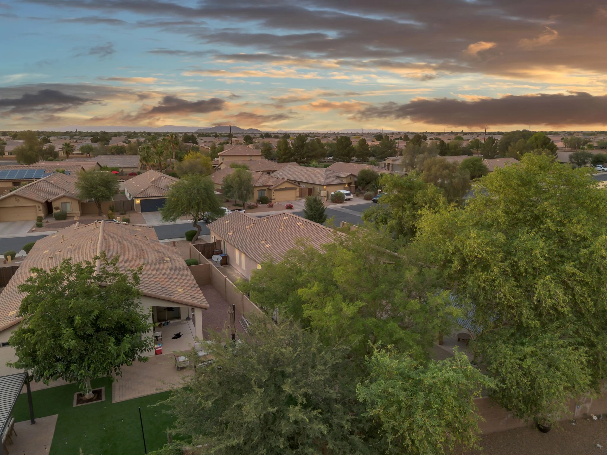 A suburban neighborhood at sunset with houses, trees, and a cloudy sky with orange and blue hues.