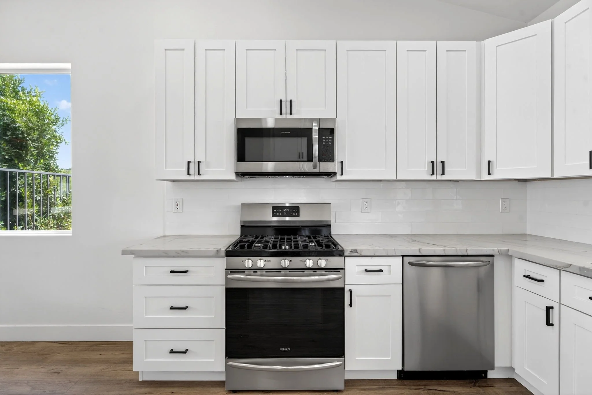 Modern kitchen with white cabinets and stainless steel appliances, including a microwave and oven, next to a window showing greenery outside.