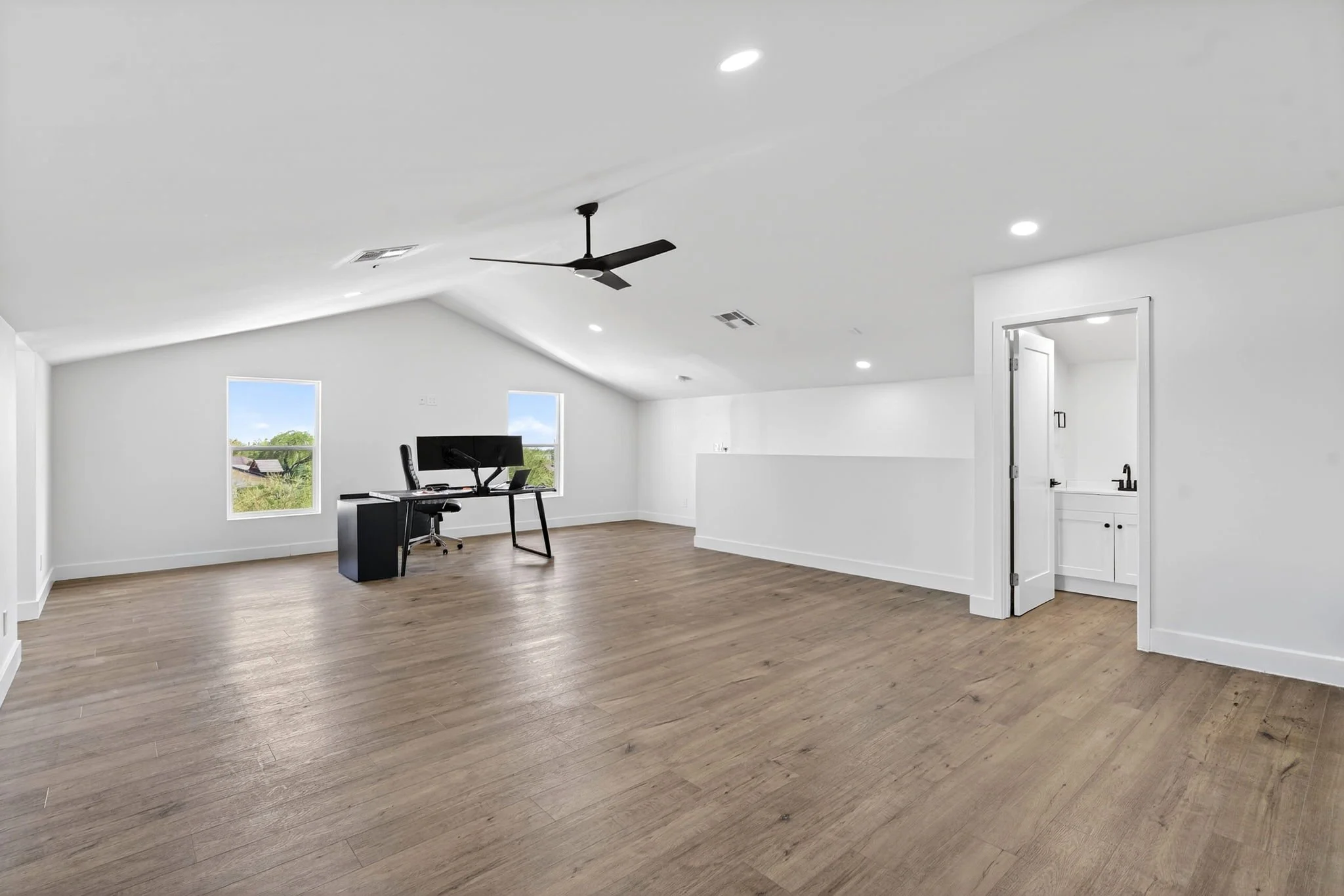 Empty room with white walls and ceiling, wood flooring, two windows showing trees outside, a desk with a computer monitor, and a ceiling fan.