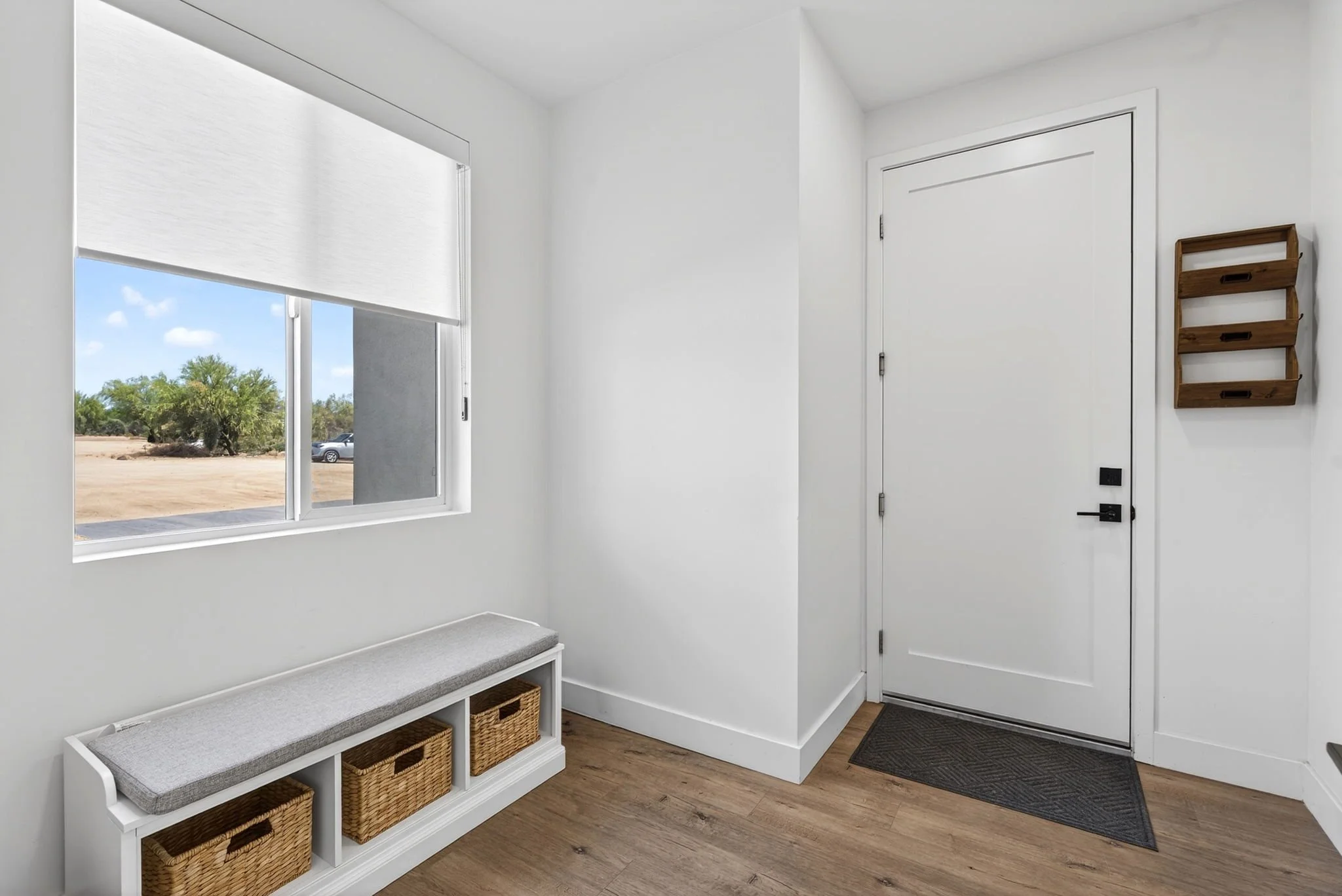 Entryway with a window, white walls, wooden flooring, a white bench with four woven baskets underneath, and a wooden wall-mounted rack with three shelves next to a closed white door