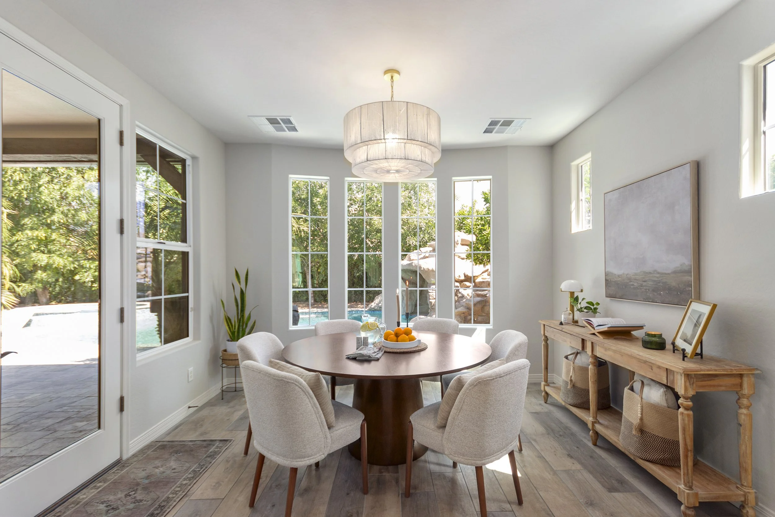 Bright dining room with large windows showing greenery outside, round wooden table with white chairs, fruit bowl, and a wooden sideboard with decor.