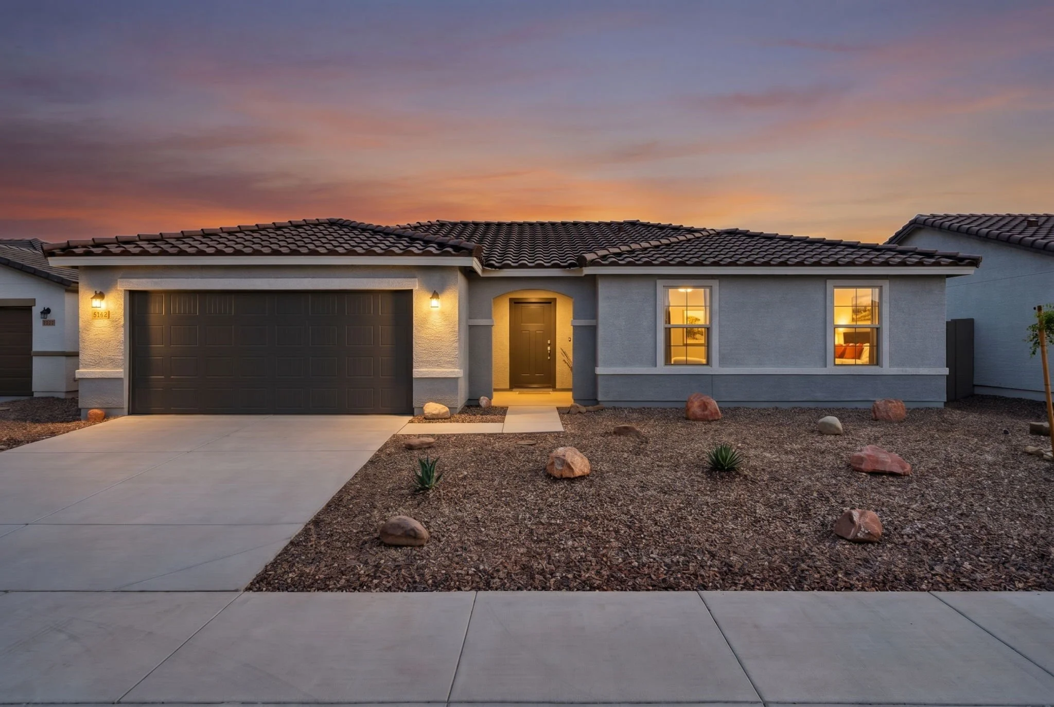 Front view of a modern single-story house at sunset, with a parking space, a pathway, windows, and exterior lighting, surrounded by a dry landscaped yard with rocks and small plants.