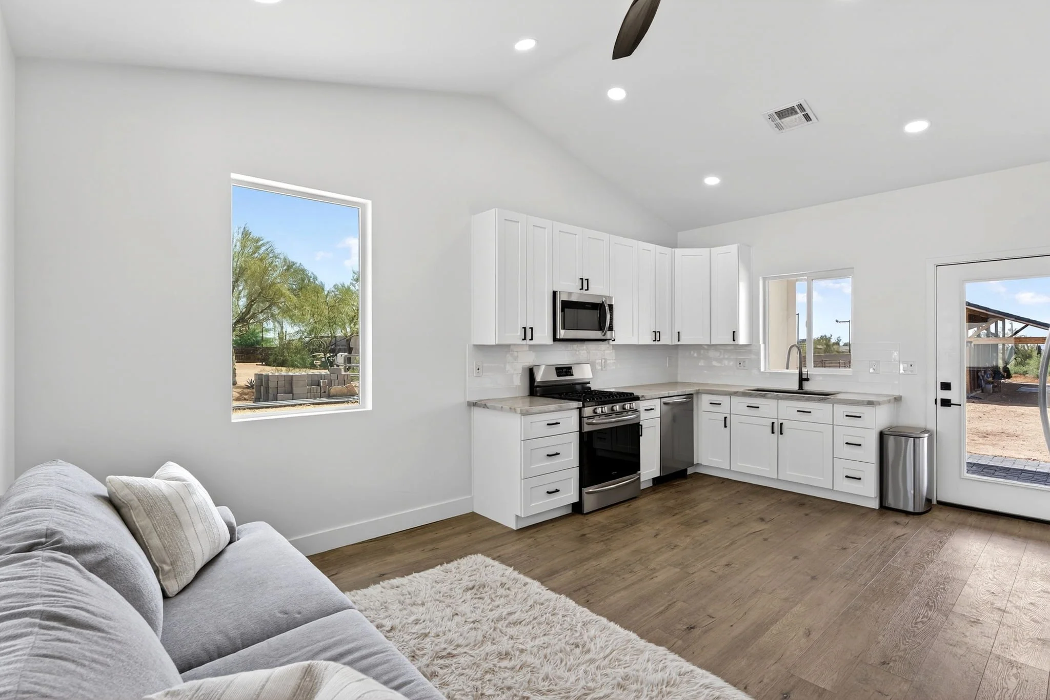 Modern kitchen with white cabinets, stainless steel appliances, and large windows showing an outdoor backyard area.