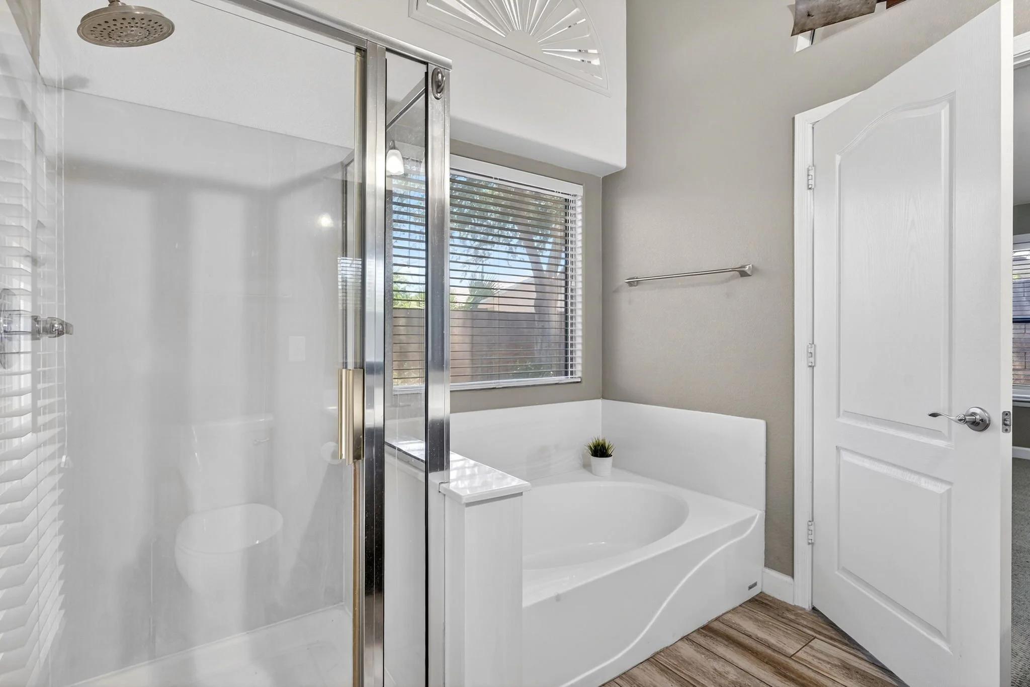 A modern bathroom featuring a white bathtub, a glass shower enclosure, a large window with blinds, and a small potted plant on the bathtub edge.