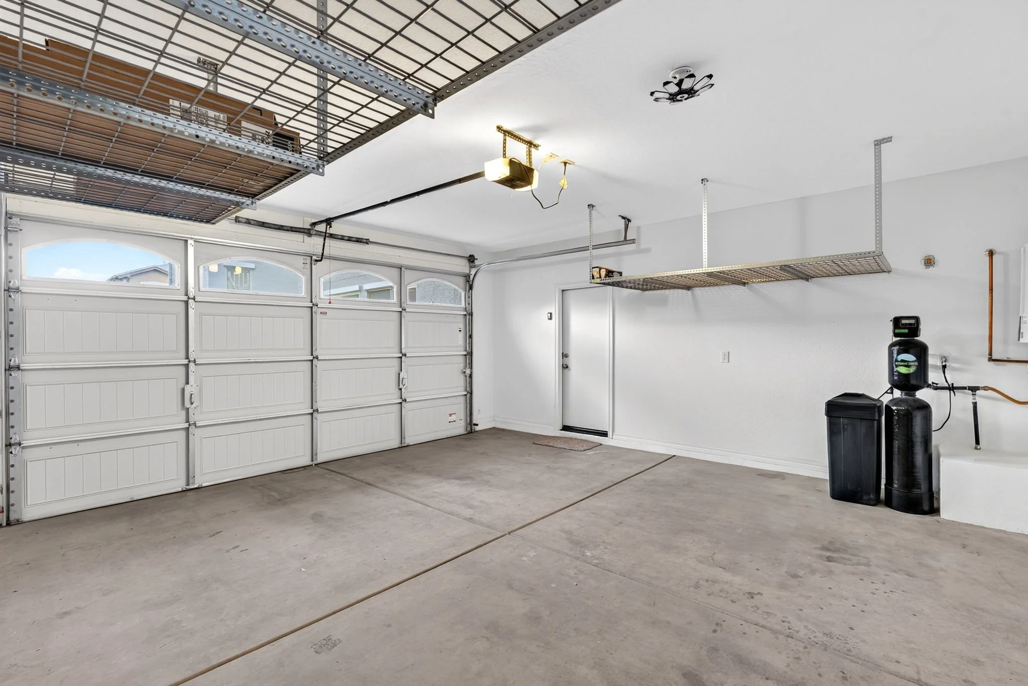 Empty garage with two overhead storage shelves, garage door, ceiling light fixture, and water softener system.