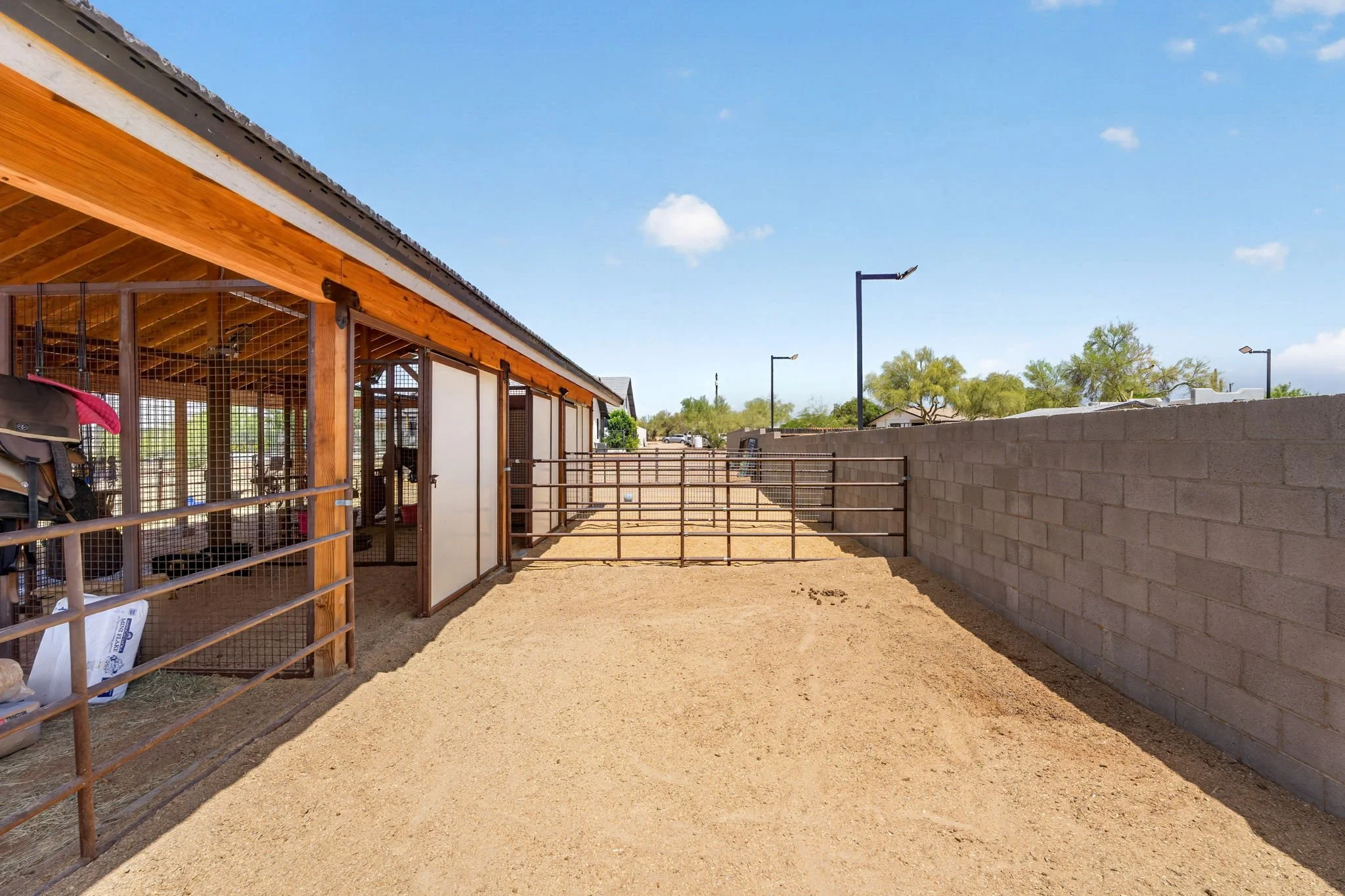 A fenced outdoor area with a building featuring a wooden roof, a concrete block wall, a dirt ground, and some trees in the background under a blue sky with a few clouds.