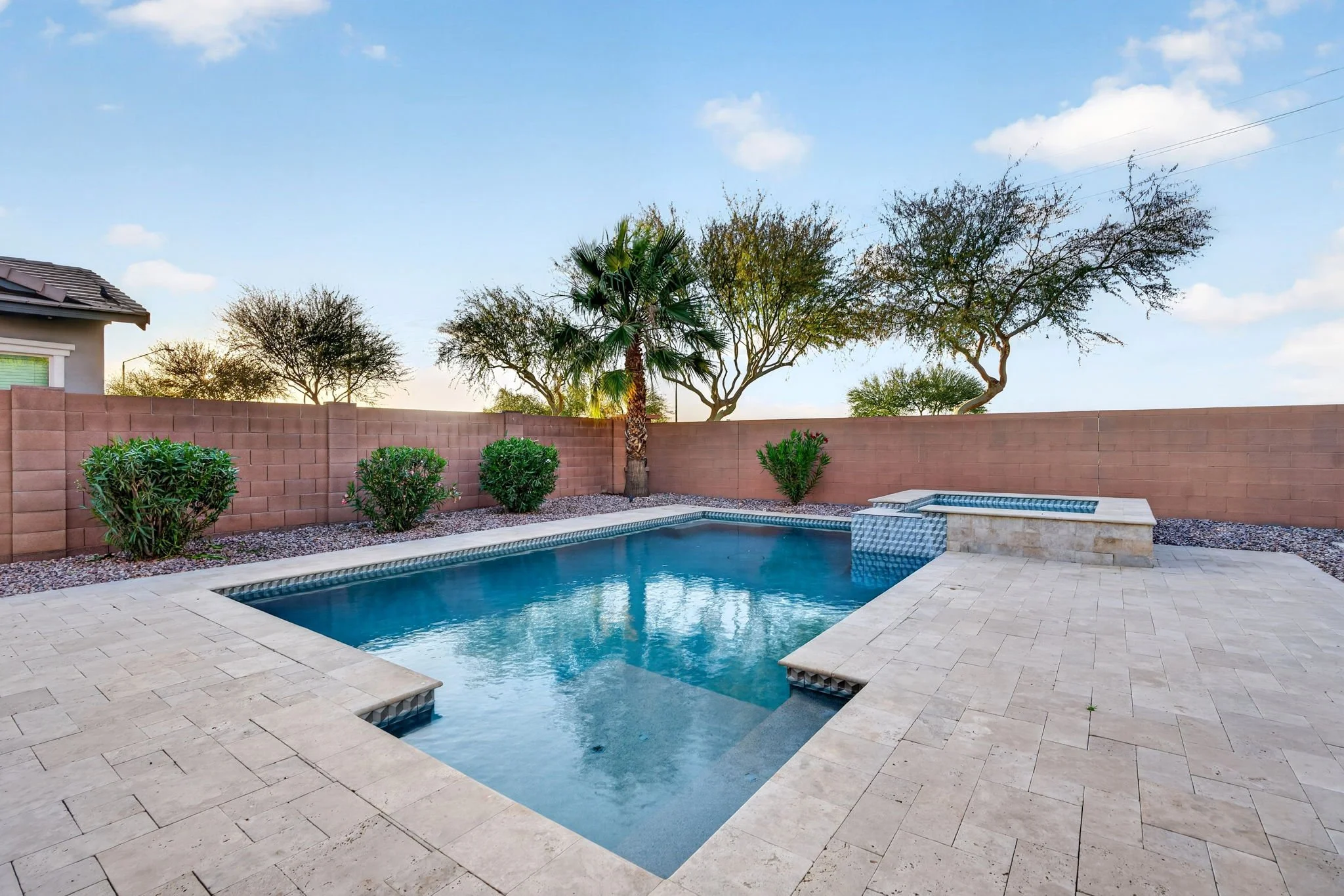 Backyard with a swimming pool, palm tree, shrubs, a high brick wall, and a clear sky with some clouds.