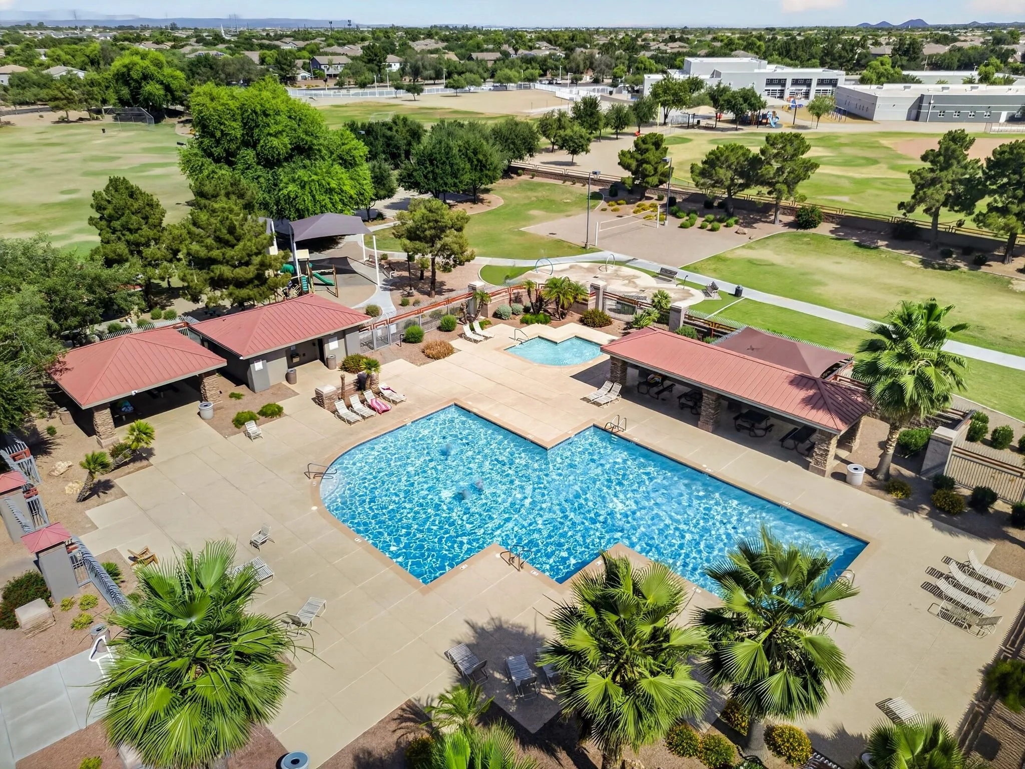Aerial view of a swimming pool area with lounge chairs, surrounded by palm trees and adjacent to a recreational facility with a playground and open grassy fields. In the background, there are residential houses and sports fields.