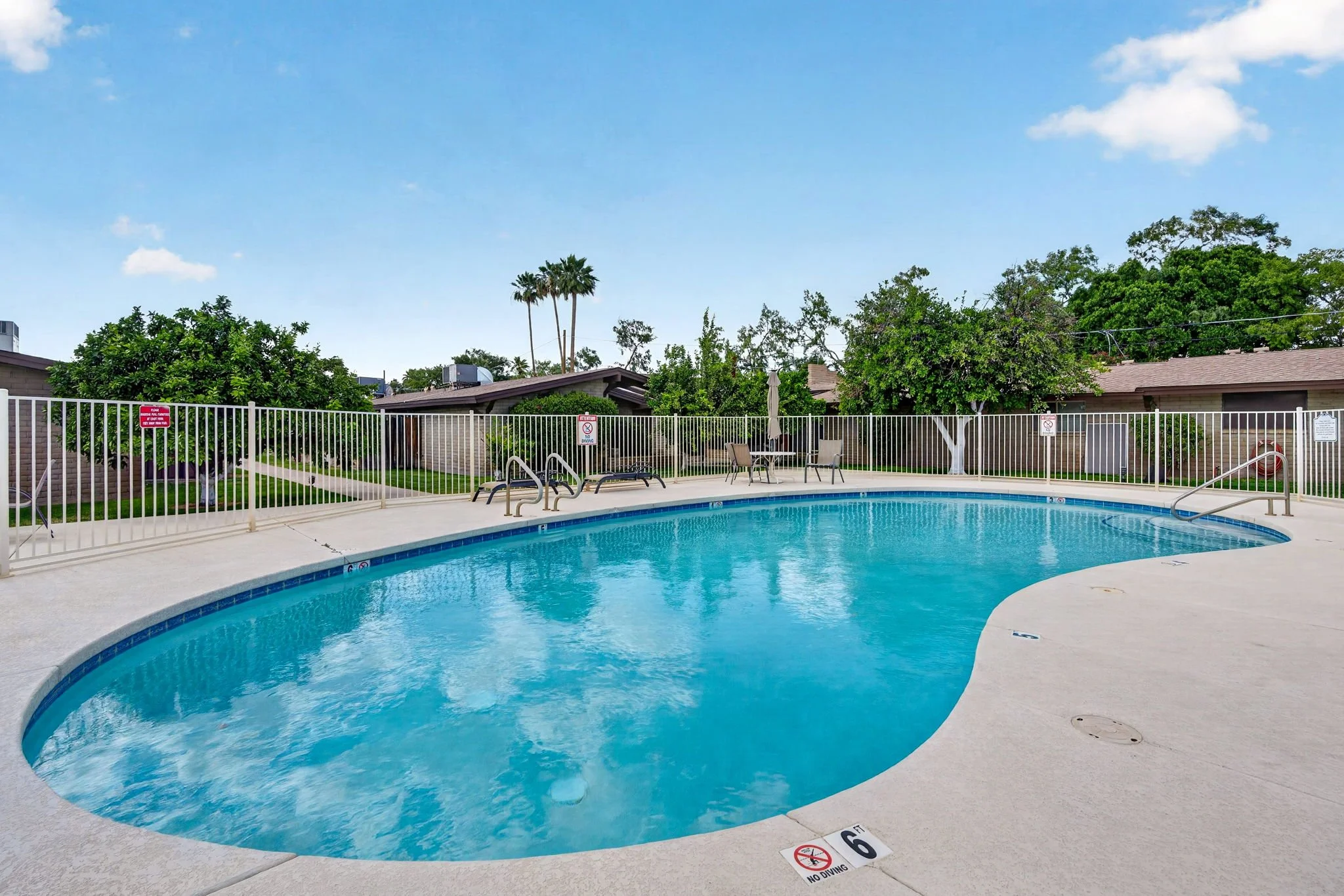 Swimming pool with blue water, surrounded by a white fence, with lounge chairs, a closed umbrella, and lush green trees and bushes in the background under a partly cloudy sky.