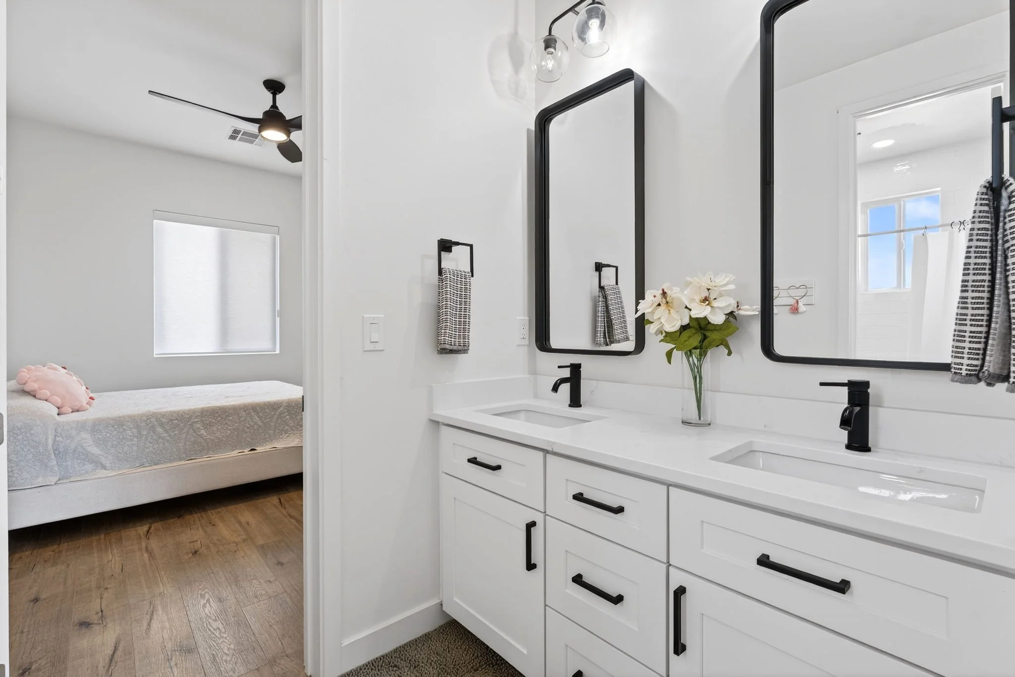 Bathroom vanity with two black-framed mirrors, black faucets, and a vase of white flowers; glimpse of bedroom with window and ceiling fan in background.
