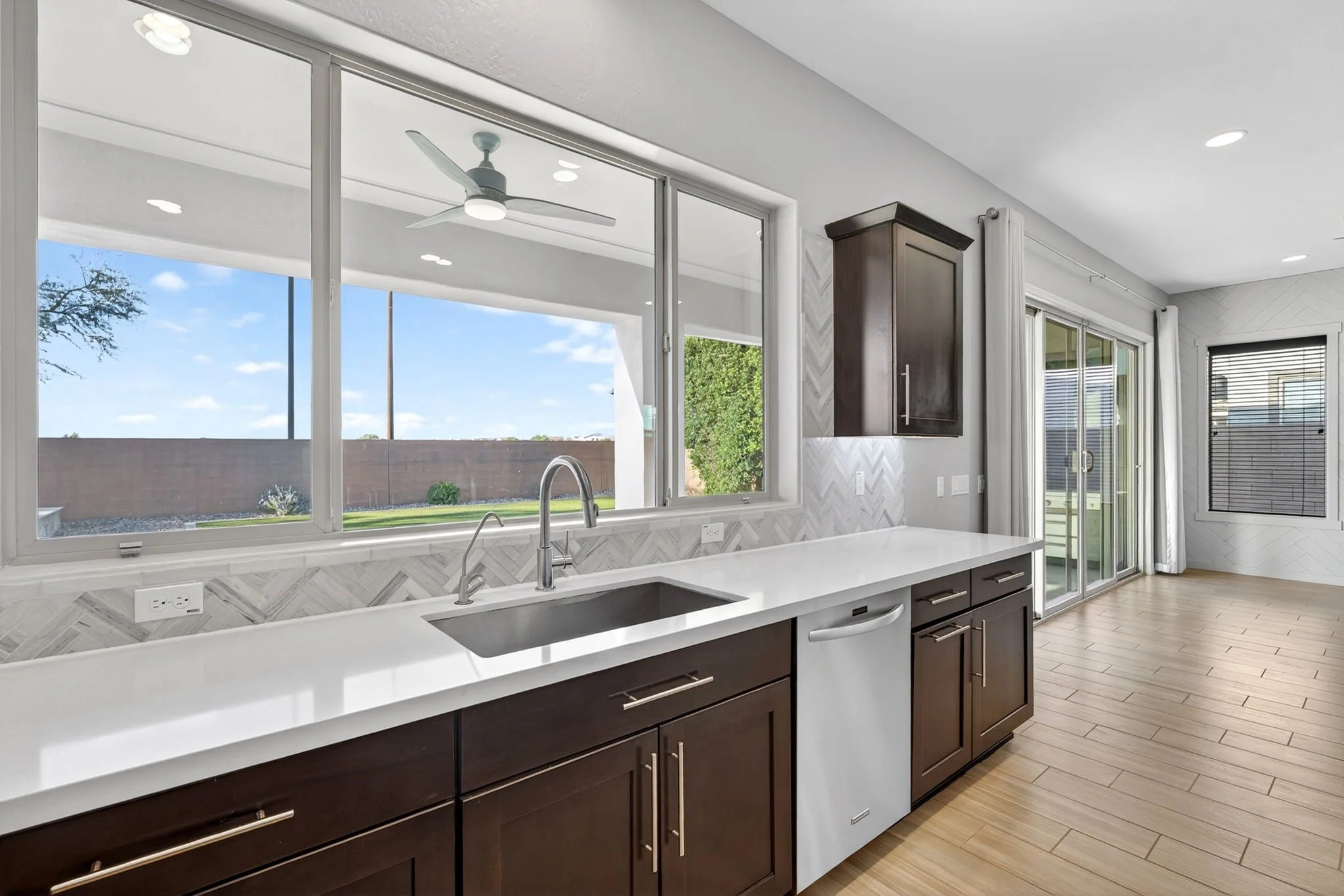 Modern kitchen with dark cabinets, white countertops, stainless steel sink, large window, sliding glass door, white ceiling, wood flooring, and ceiling fan outside.