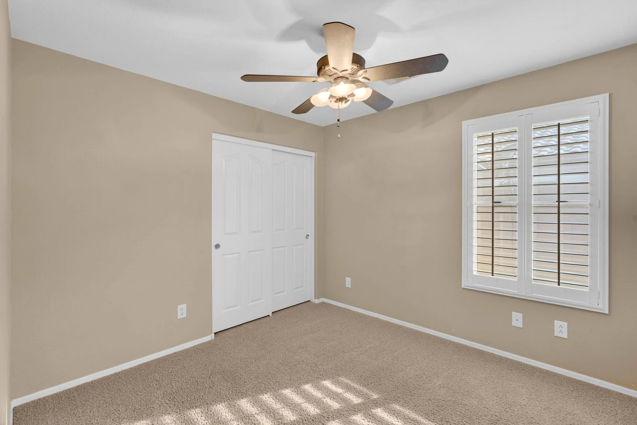 Empty beige room with white trim, carpeted floor, two windows with blinds, ceiling fan with lights, and a closet with double doors.