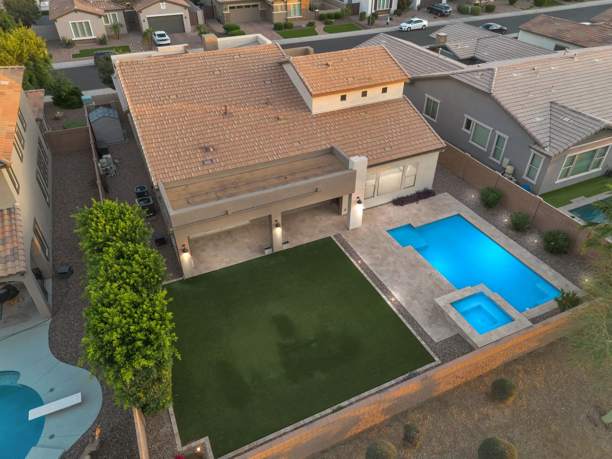 Aerial view of a residential backyard with a swimming pool, a patio, a lawn, and neighboring houses with tiled roofs.