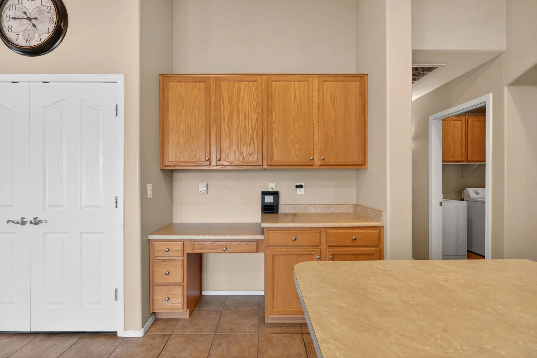 Kitchen with light wood cabinets, beige countertops, and a wall clock showing 9:08, with a laundry room visible through an open doorway to the laundry area.