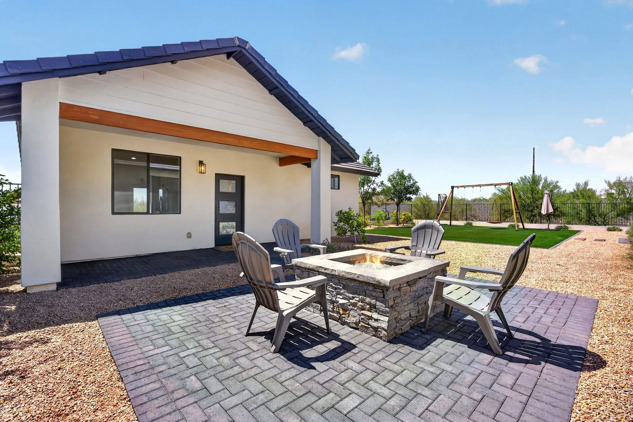 A backyard with a house, a stone fire pit with chairs around it, a swing set, and a fence with trees in the background under a blue sky.