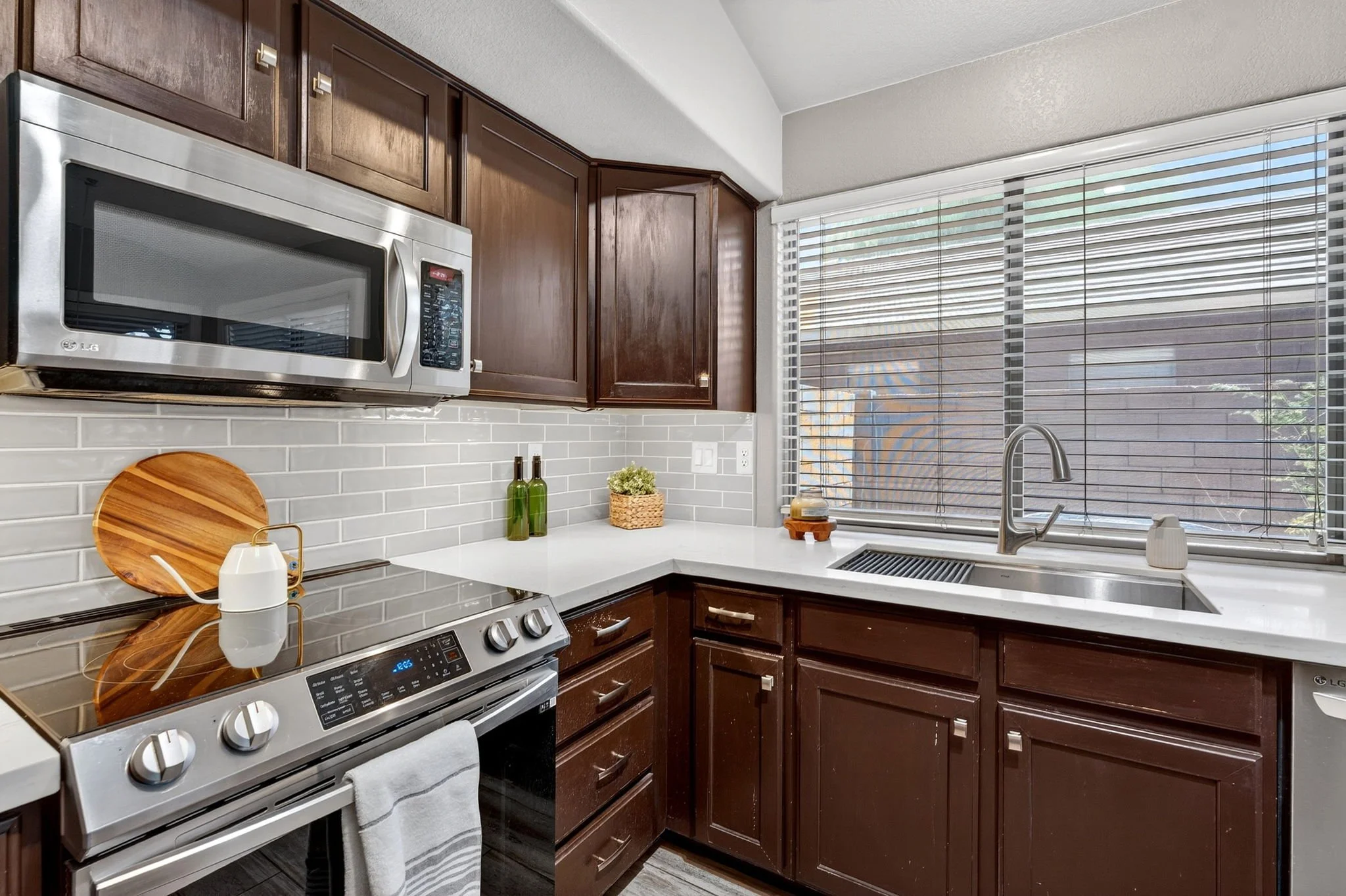 A kitchen with dark wood cabinets, stainless steel microwave and stove, white countertops, and a large window with blinds. Items on the counter include a wooden cutting board, two green bottles, a small plant, and some soap and dish items near the si