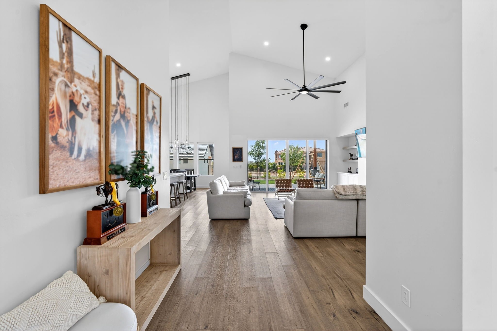 Open living room with white walls, wooden flooring, and a large ceiling fan. There are two white sofas, a wooden console table with framed awards and small plant, wall art, and a view of the backyard through large glass doors.