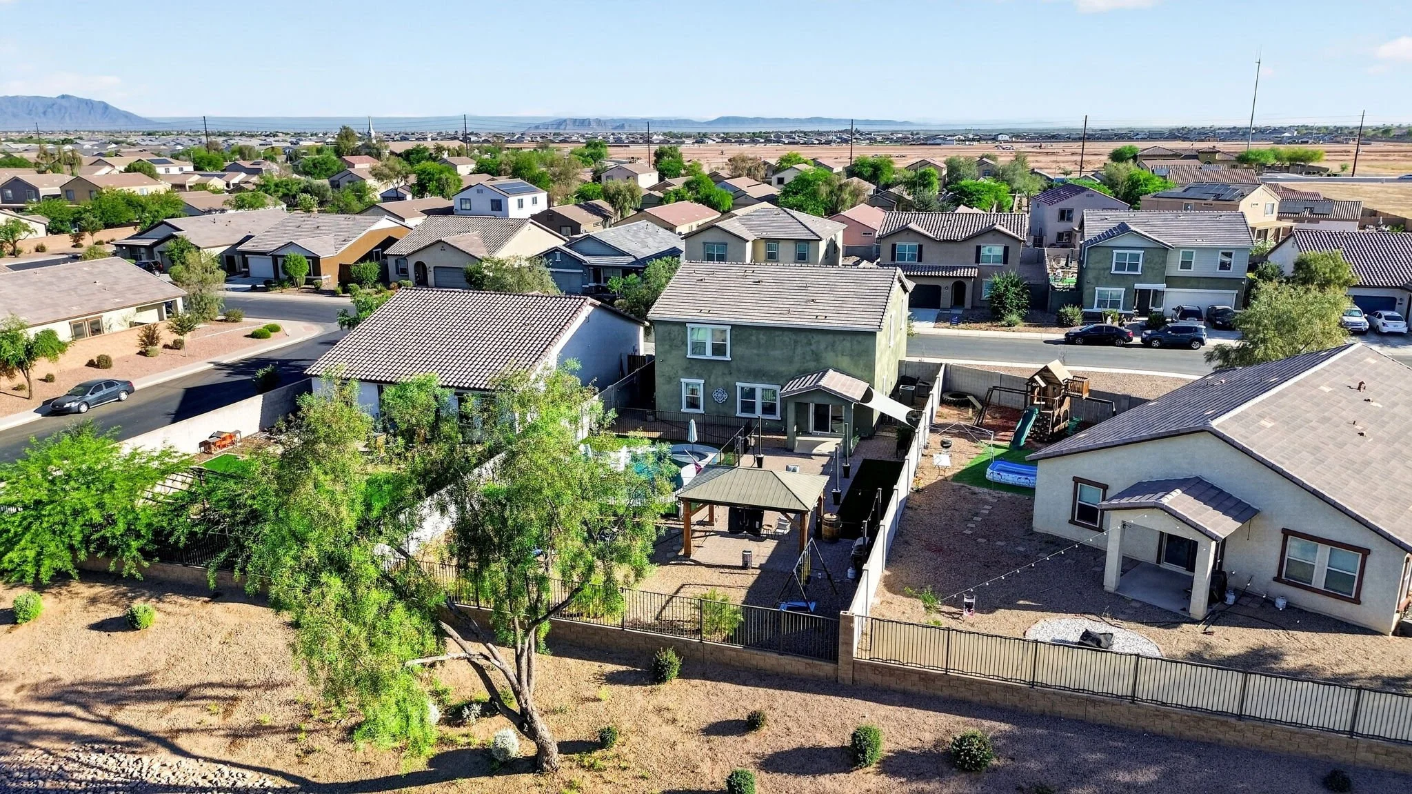 A suburban neighborhood with houses, trees, and a backyard with a patio and children's play area, with hills and mountains in the background.