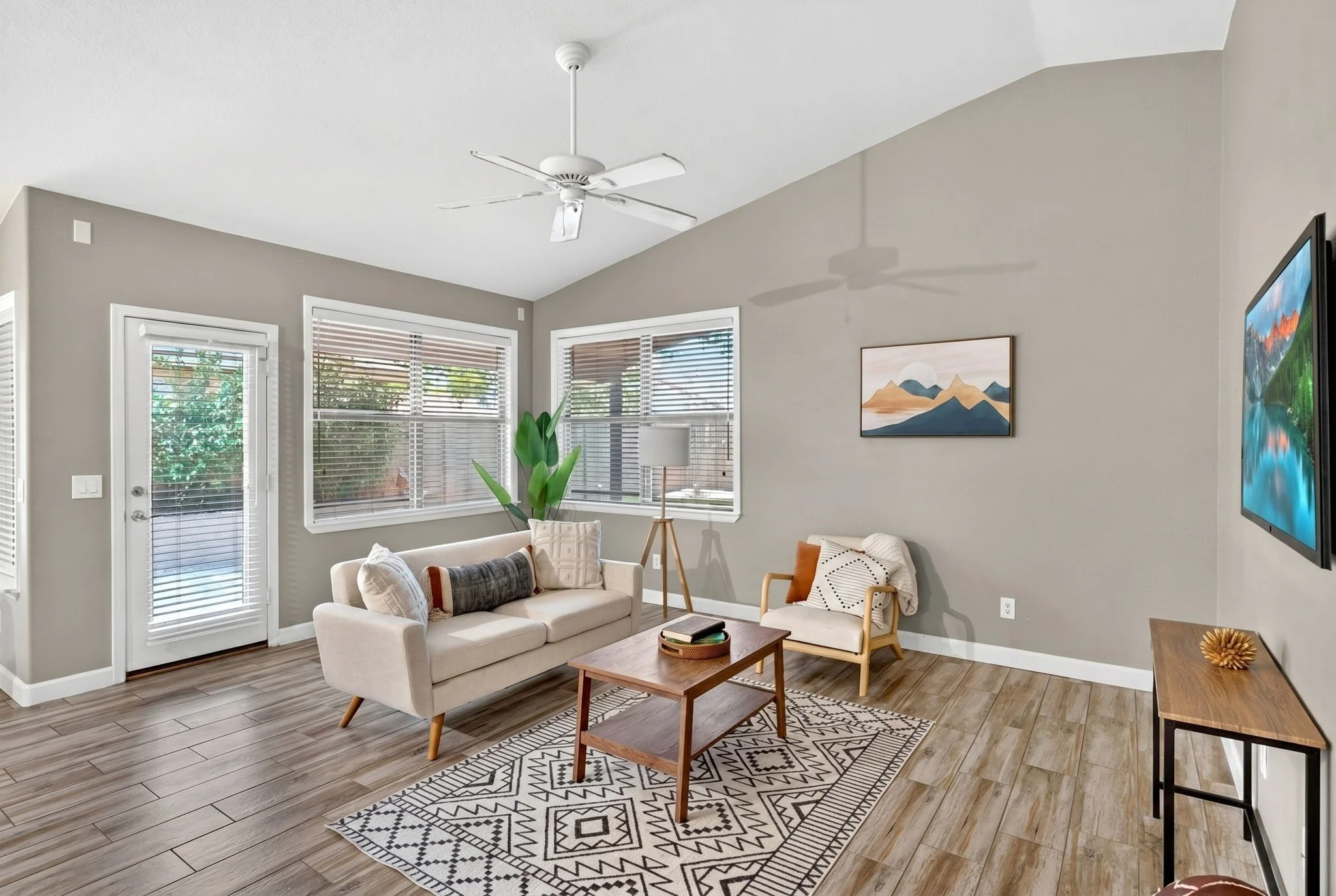 Living room with large windows, a white sofa, a wooden coffee table, a patterned rug, a potted plant, wall art, and a flat-screen TV on the wall.