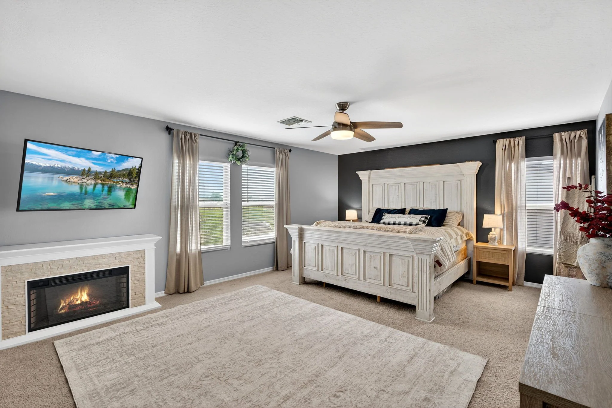 A bedroom with a white bed frame, beige bedding, navy pillows, a bedside lamp, and windows with beige curtains. There is a gray accent wall, a ceiling fan, a wall-mounted TV showing a lake scene, and a fireplace with a fire.
