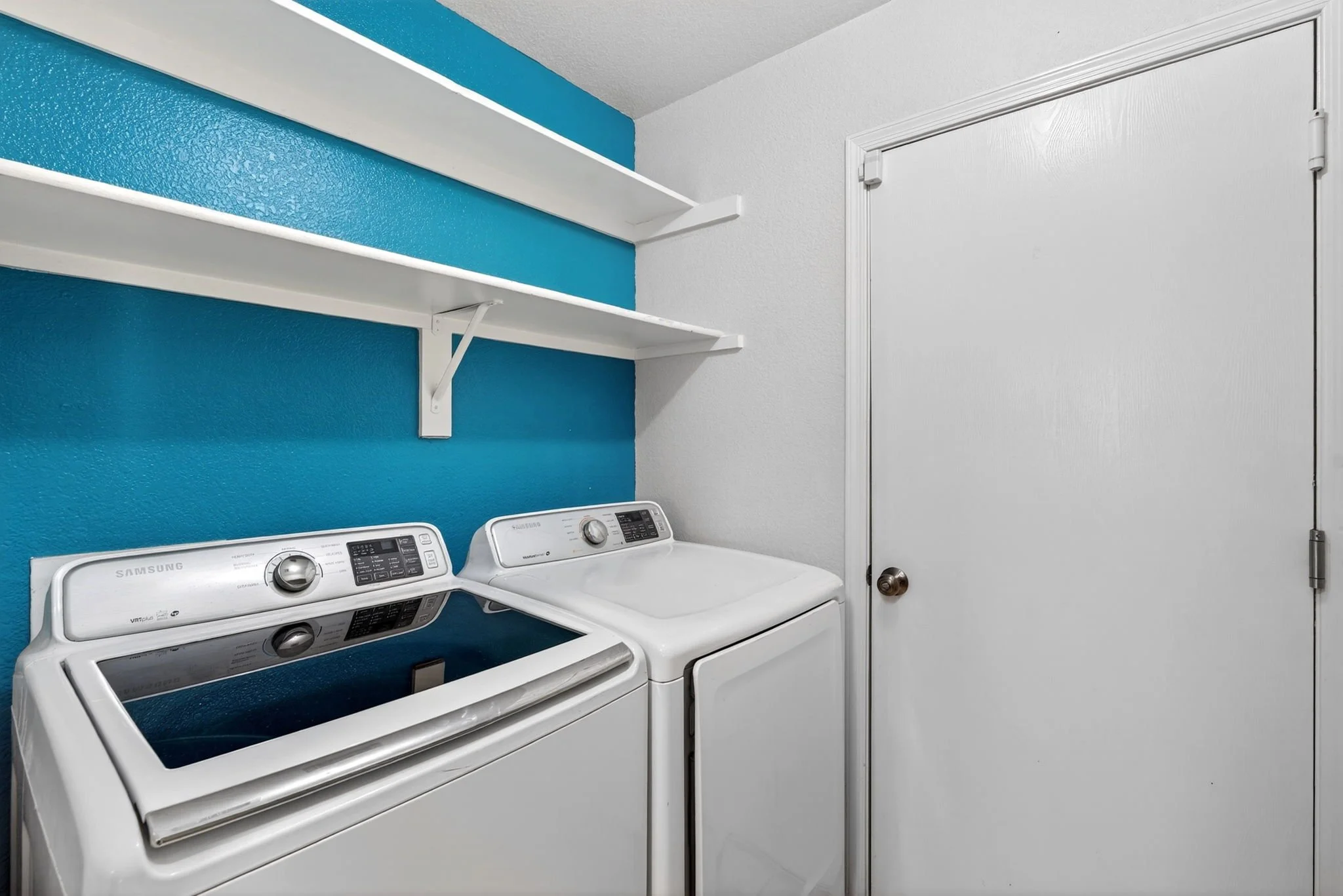 A laundry room with a white washing machine and dryer, a blue wall behind them, a white shelf above, and a closed white door to the right.