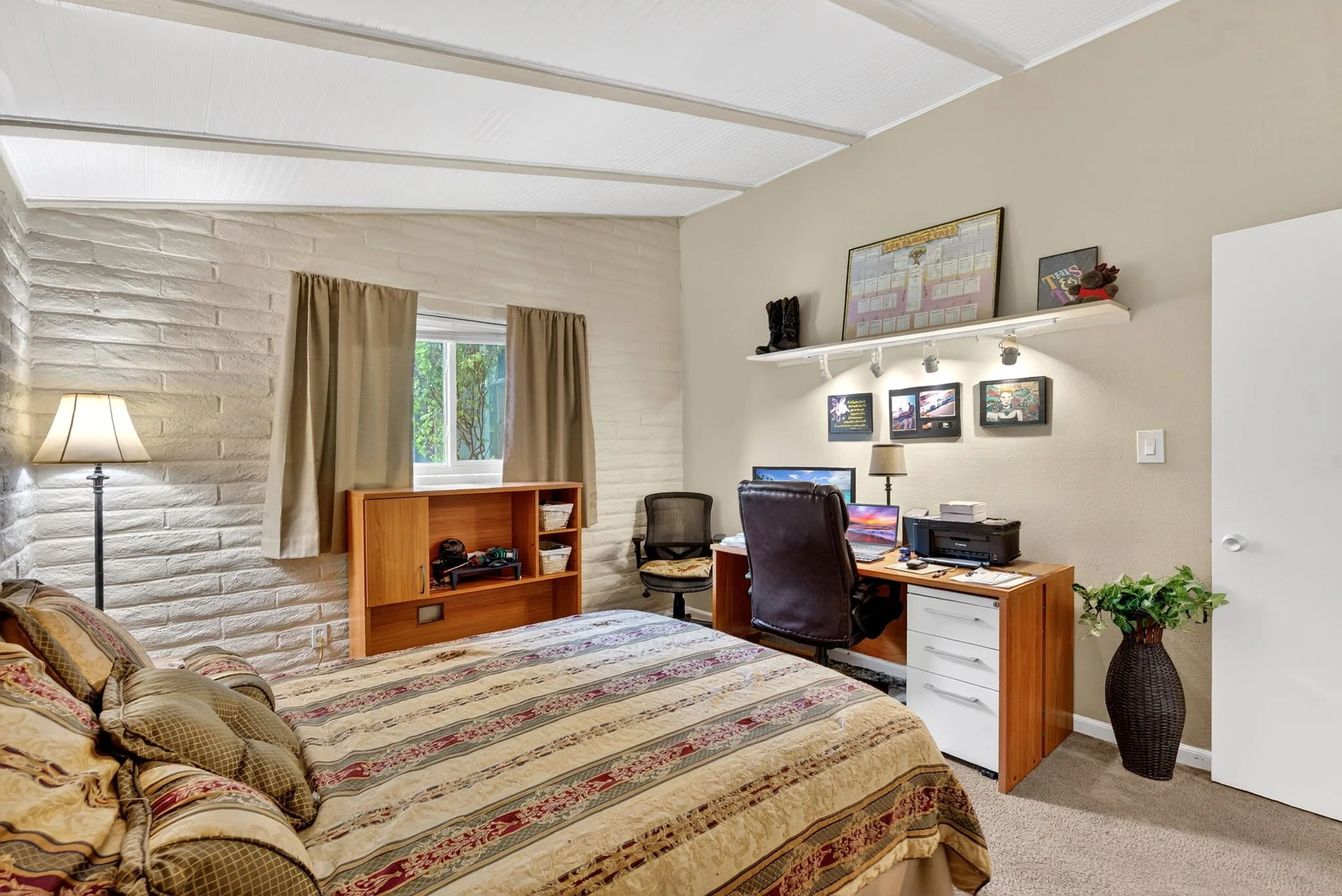 A bedroom with a striped bedspread, a wooden nightstand, a desk with a computer, a black office chair, beige curtains on a window, a tall floor lamp, framed pictures on the wall, and a white shelf with decorative items.