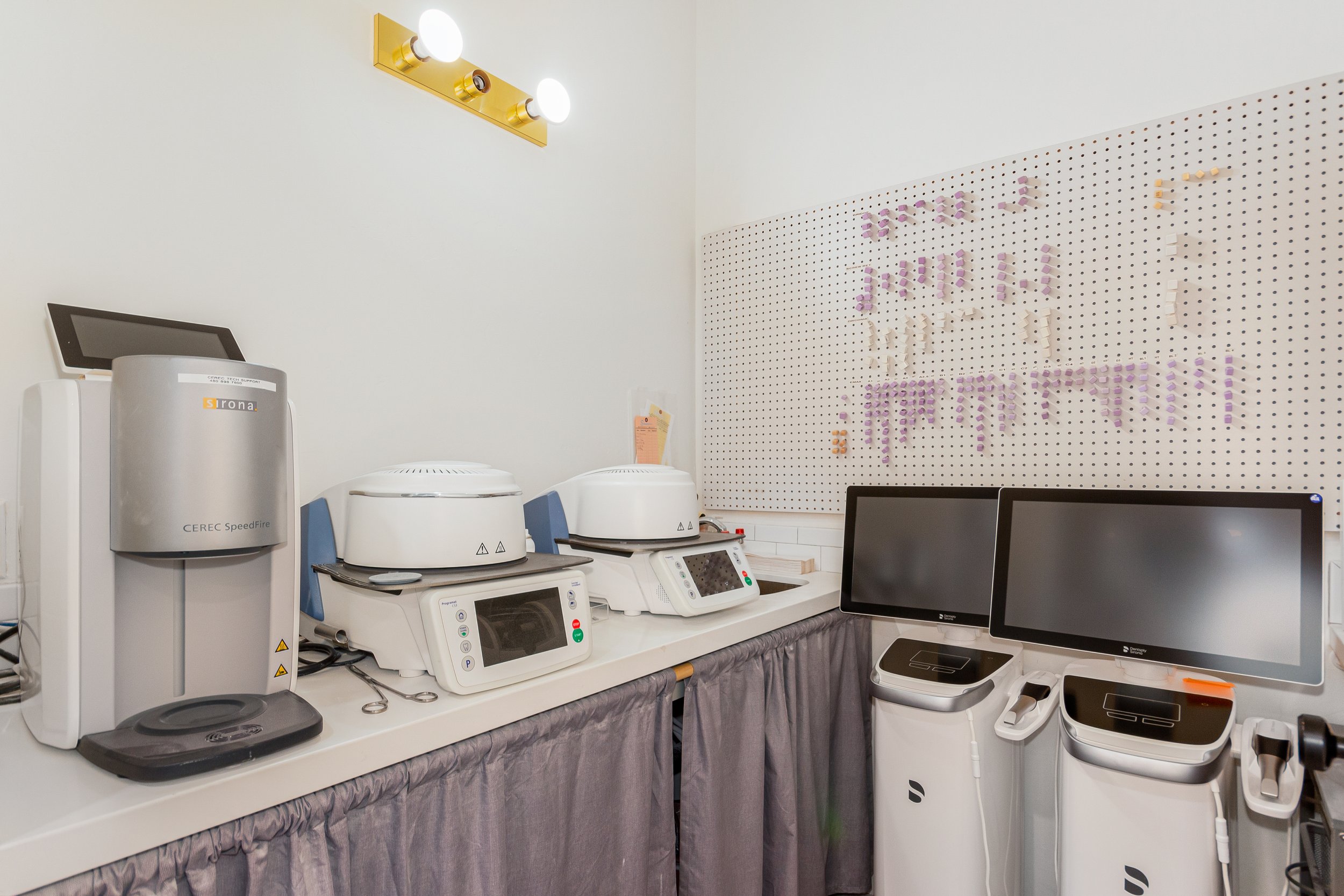 Medical or laboratory equipment on a white counter, including centrifuge machines, monitors, and other devices, with a white pegboard wall with purple and yellow clips and a light fixture on the wall.