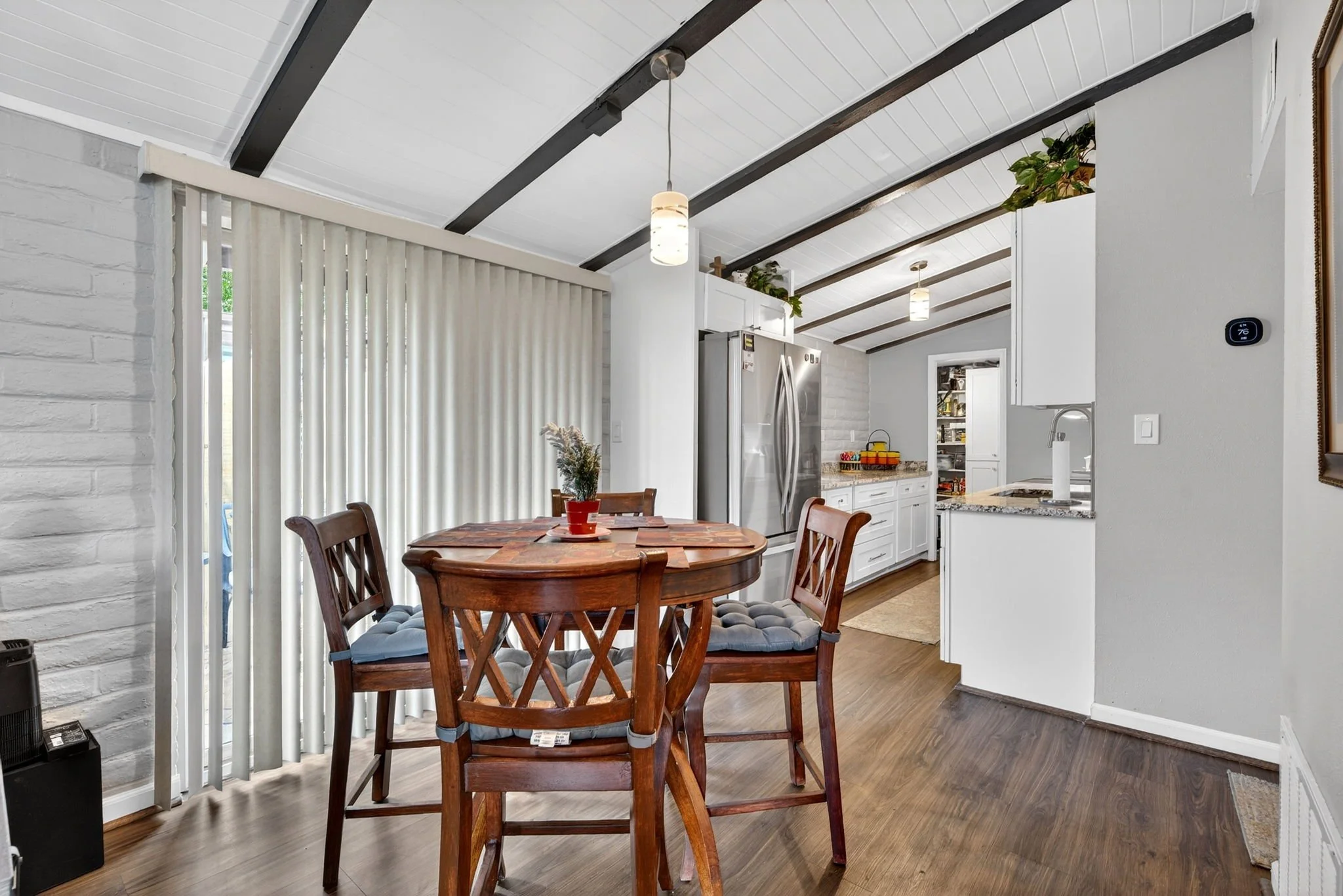 A dining area with a wooden round table, four matching chairs with cushioned seats, a potted plant on the table, and sliding vertical blinds covering a sliding door. The background shows a kitchen with white cabinets, stainless steel refrigerator, an