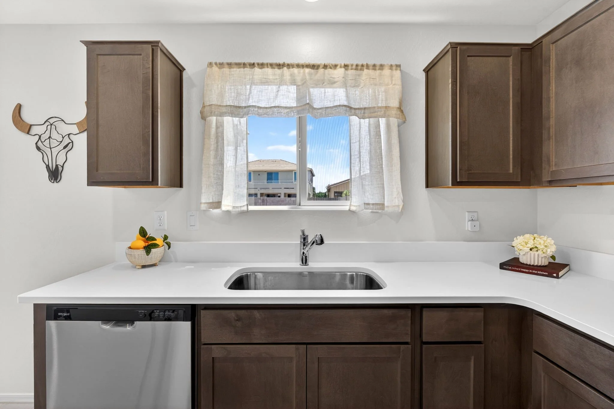 Kitchen with white countertop, stainless steel sink, wooden cabinets, window with beige curtains, decorative bull skull wall art, fruit bowl, and flower pot.