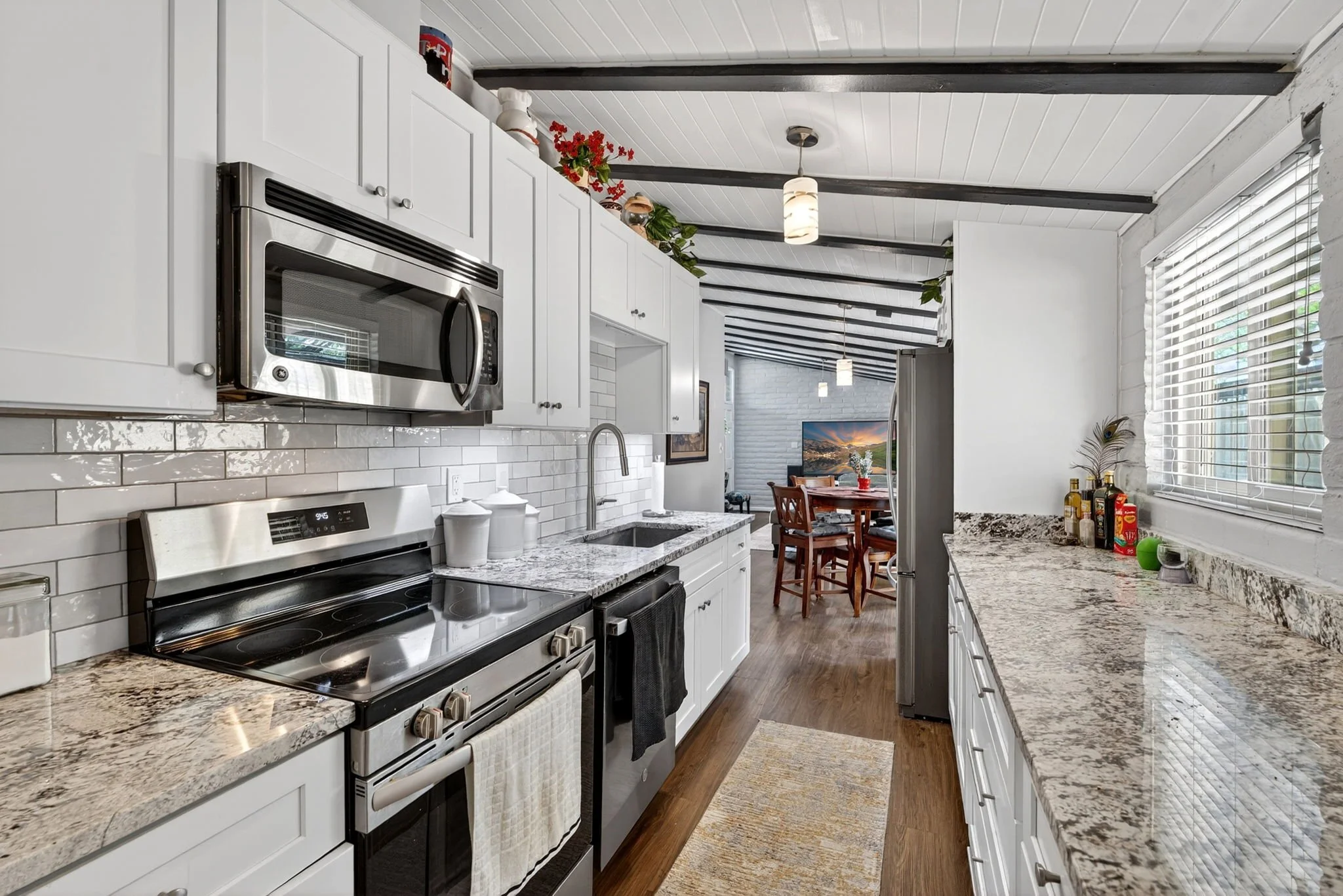 A modern kitchen with white cabinets, a granite countertop, a stainless steel microwave, and a stove. There are decorative items, plants, and kitchen essentials on the counter, with a view into a dining area with a wooden table and chairs, and a TV i