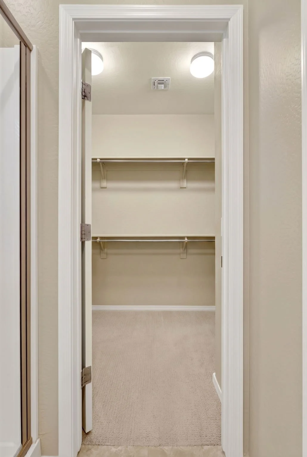 Empty walk-in closet with beige walls, white shelves, and a beige carpeted floor.
