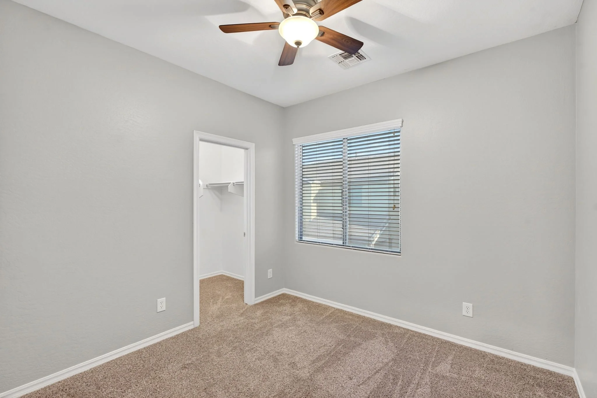 Empty room with beige carpet, light gray walls, a window with blinds, an open closet with a shelf and hanging rod, a ceiling fan with light, and multiple electrical outlets.