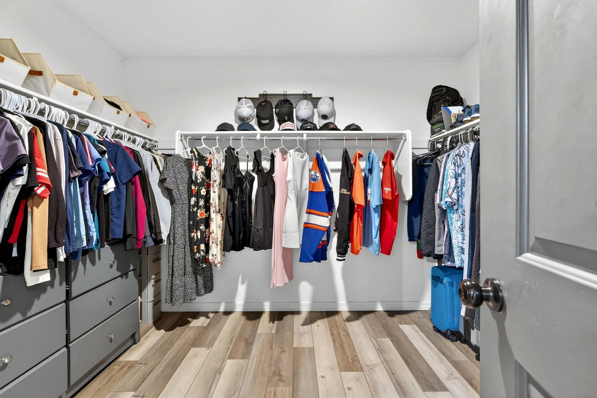 A walk-in closet with hanging clothes, hats on a shelf, and two suitcases on the floor near the door.