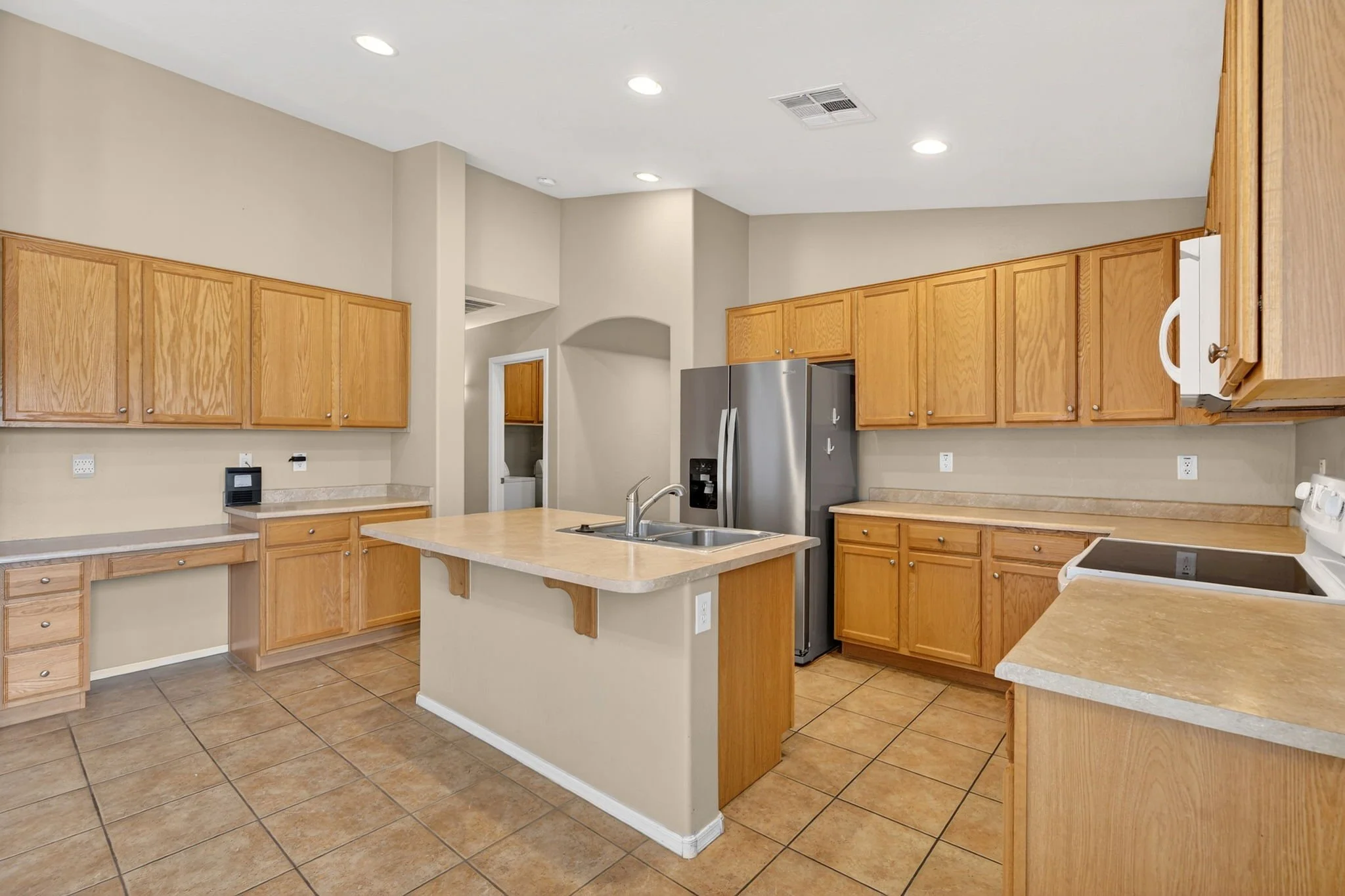 A spacious kitchen with wooden cabinets, beige countertops, and tiled floor. Contains a stainless steel refrigerator, a white stove, and a double sink. Recessed ceiling lights illuminate the space.