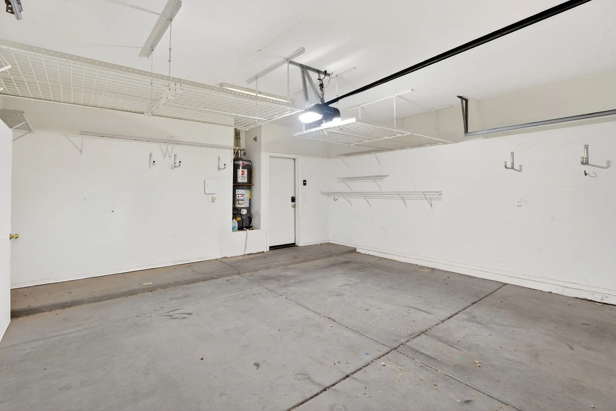 Empty garage with a concrete floor, white walls, ceiling hooks, and wire shelves on the wall. Fluorescent lights installed on the ceiling. Fire extinguisher and some tools in a small closet.