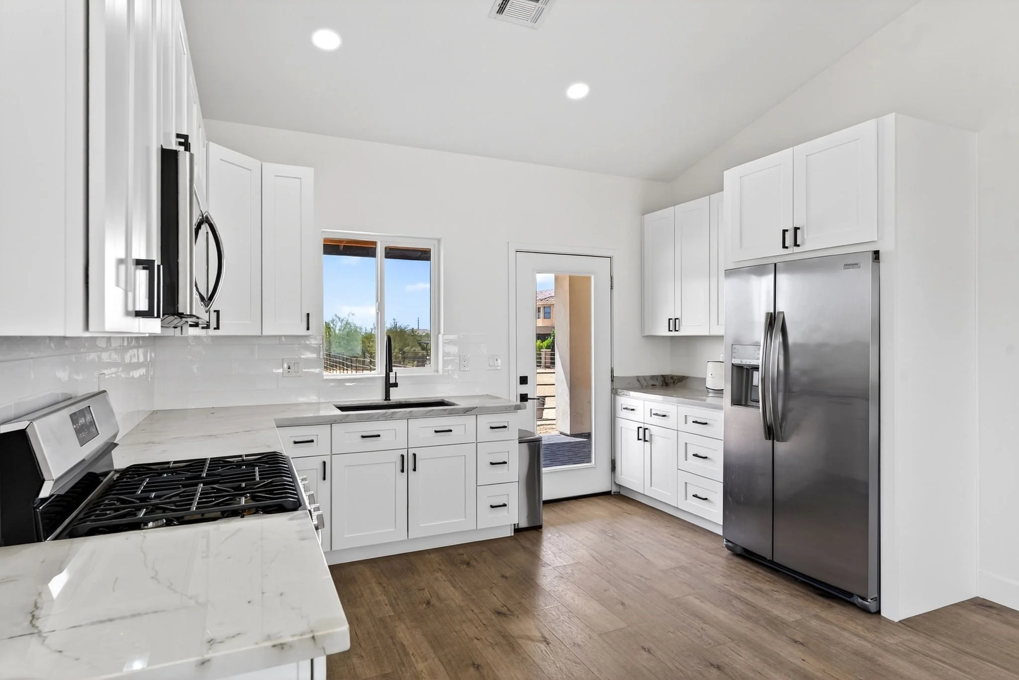 Bright modern kitchen with white cabinets, stainless steel appliances, and hardwood floors, with windows and a door leading outside.
