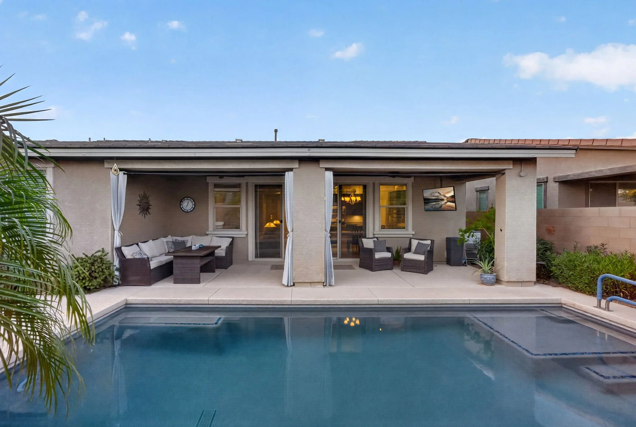 Photo of a backyard patio area with outdoor furniture, a swimming pool, and a house with a covered porch. The house has beige stucco walls, large windows, and a flat roof, with trees and plants around.