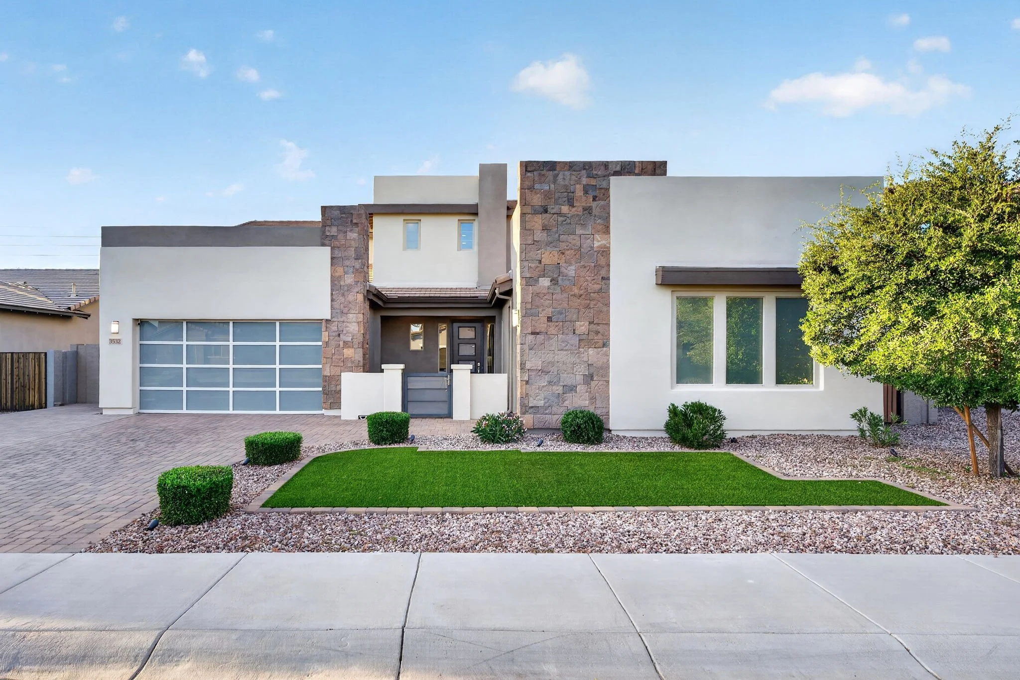 Modern suburban house with white walls, a stone accent wall, large windows, a garage door, front pathway, neatly manicured lawn, small bushes, and a tree under a blue sky with clouds.