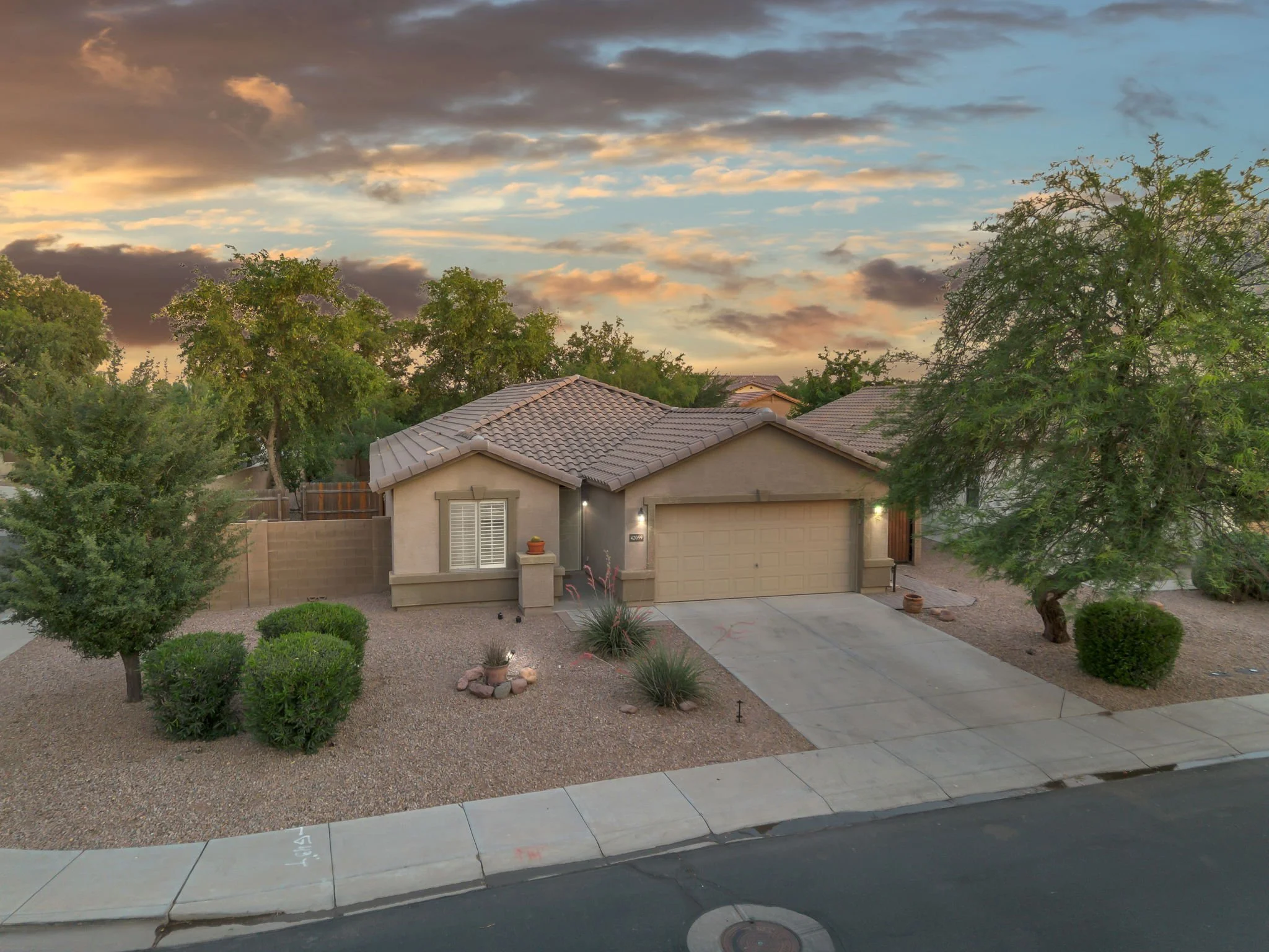 Residential home with tiled roof, front yard with gravel, bushes, trees, and a sidewalk under a partly cloudy sky at sunset.