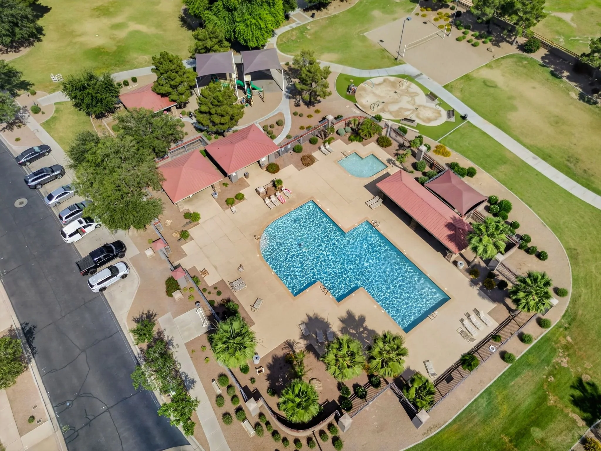 Aerial view of a community park with swimming pools, a playground, picnic tables, parking lot, green lawns, trees, and walking paths.