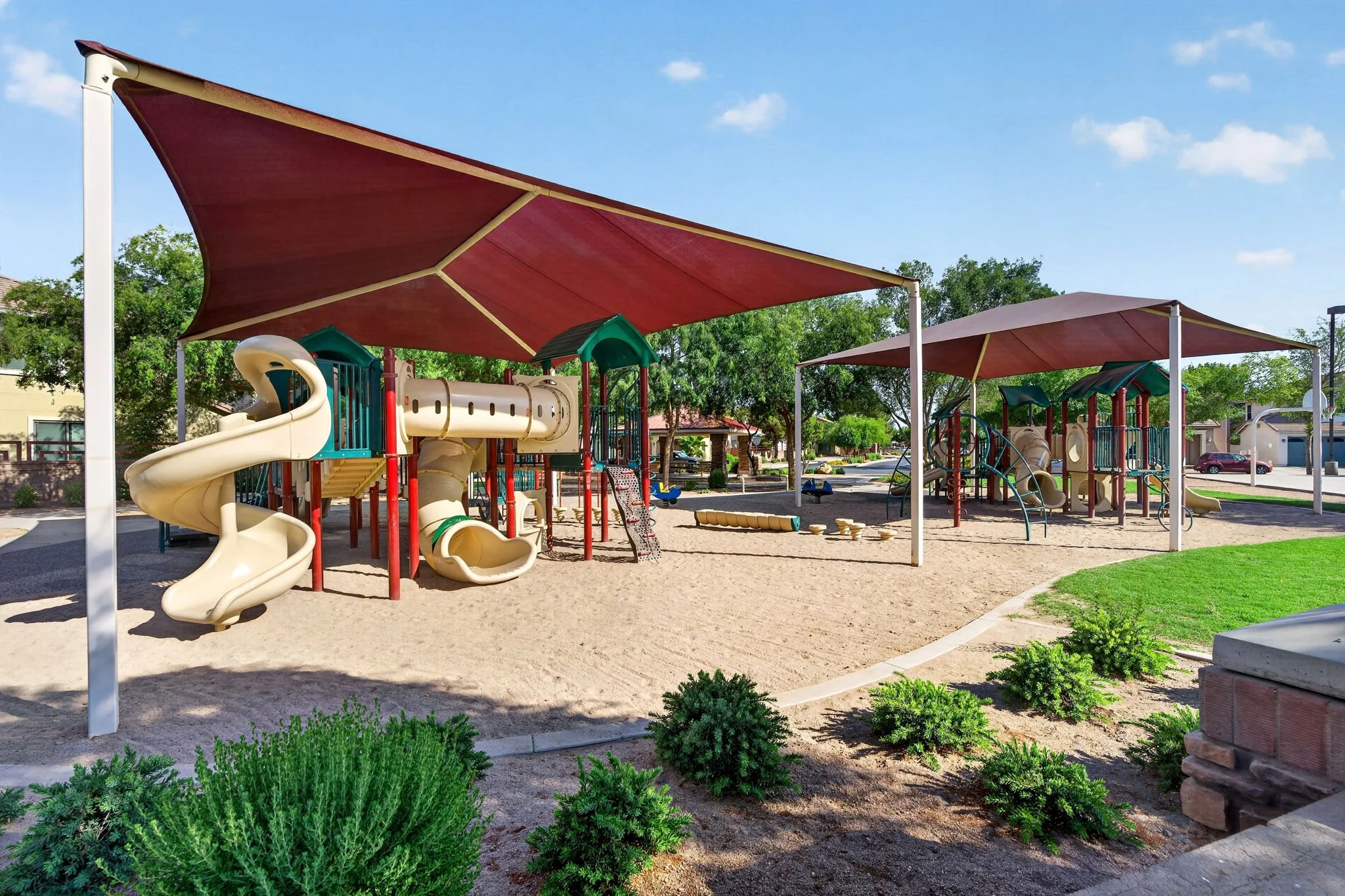 A playground covered by large red shade sails with slides and climbing structures, surrounded by trees, green bushes, and a paved walkway in a sunny outdoor park.