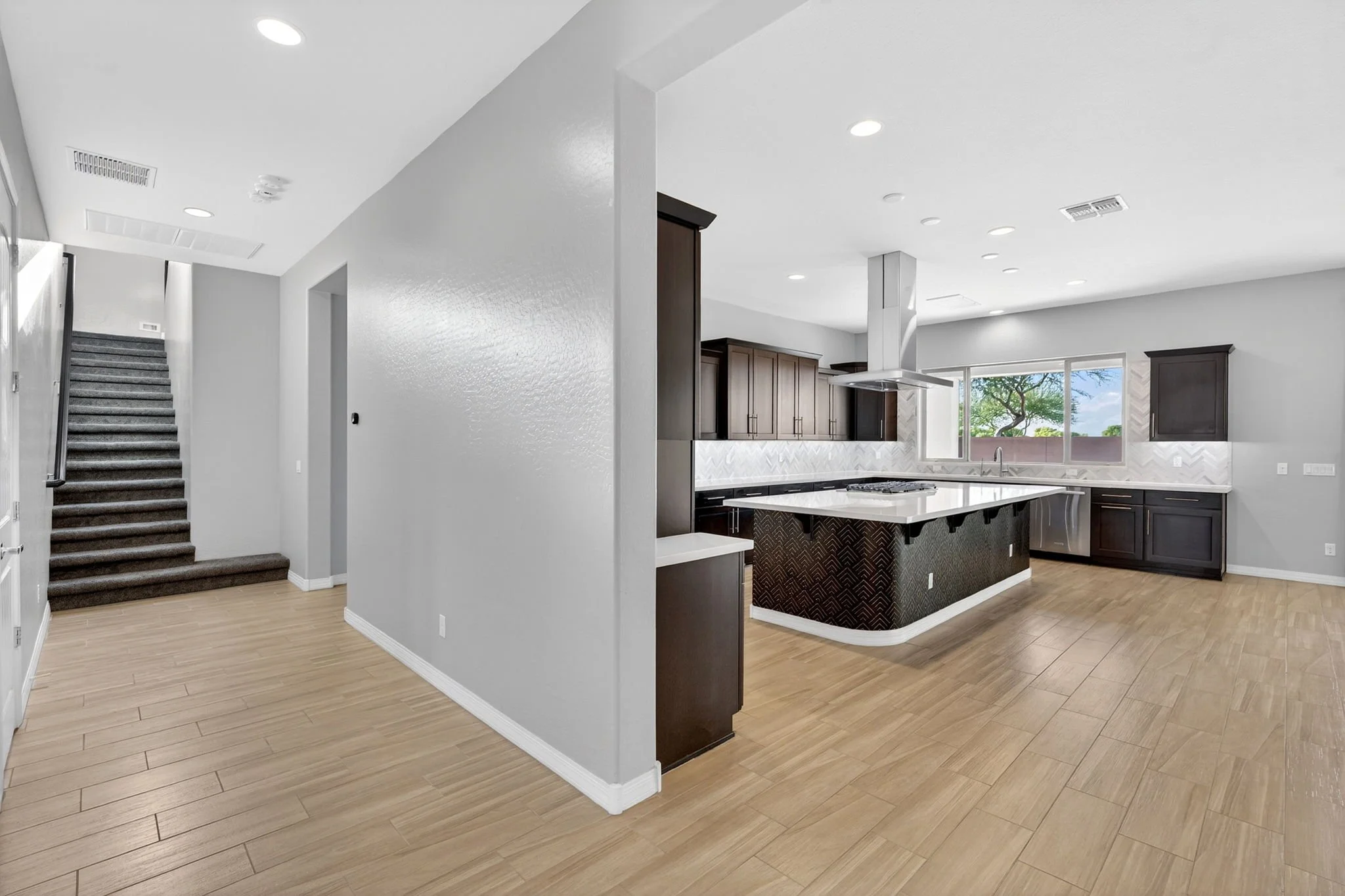 Open-concept living space with kitchen featuring dark wood cabinets, a large white island, and modern appliances. Light wood flooring and large window showing greenery outside. Stairs with carpeted steps are visible to the left.