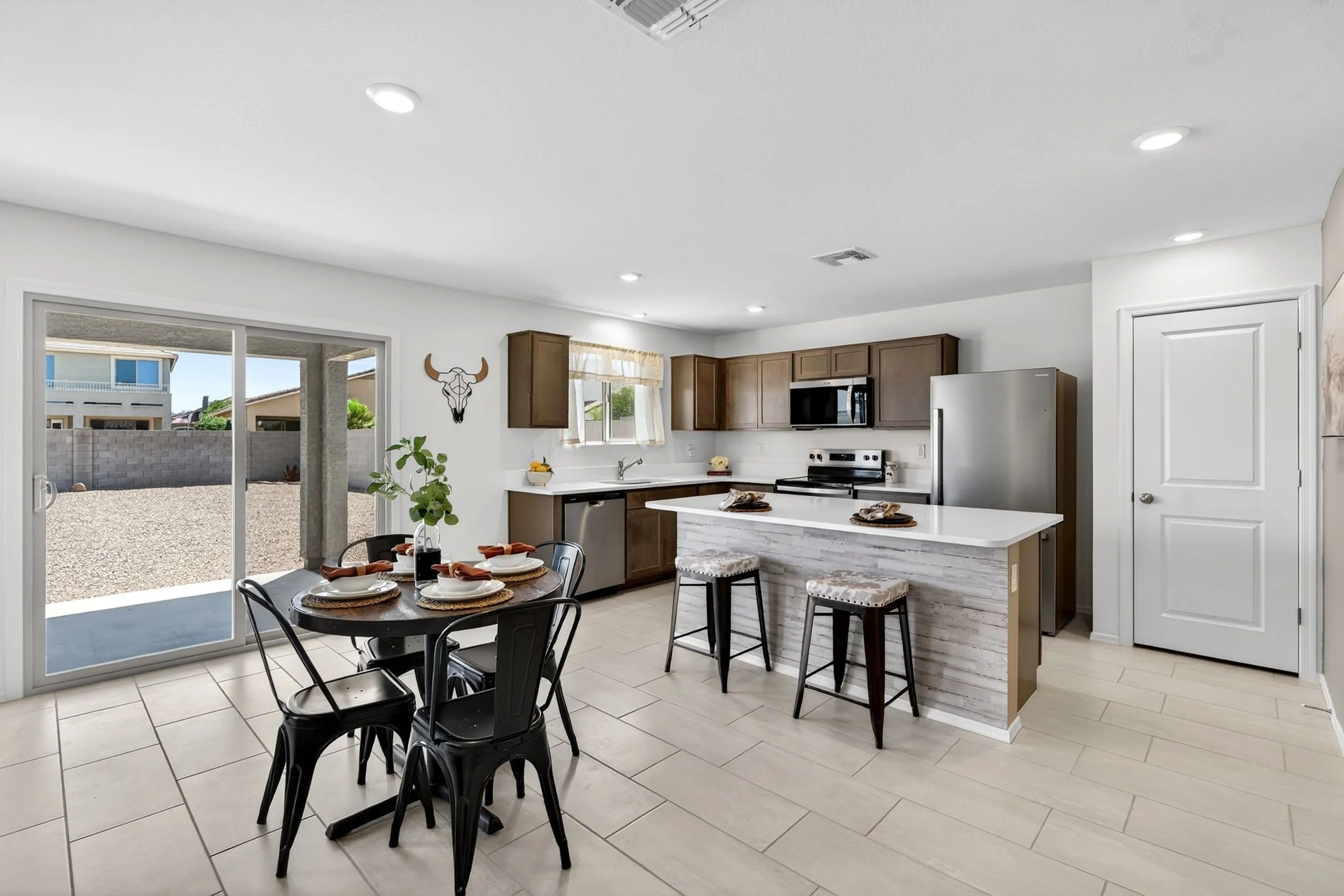 Open-concept kitchen and dining area with sliding glass door leading to the backyard, featuring a round dining table set with plates, napkins, and a centerpiece, alongside wooden cabinets, stainless steel appliances, and a decorative bull skull wall 