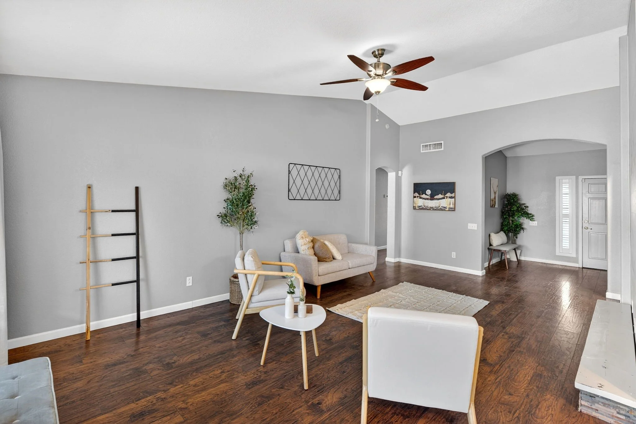 Living room with light gray walls, wooden floors, ceiling fan, beige sofa, white chairs, small coffee table with vases, artwork, and indoor plants.