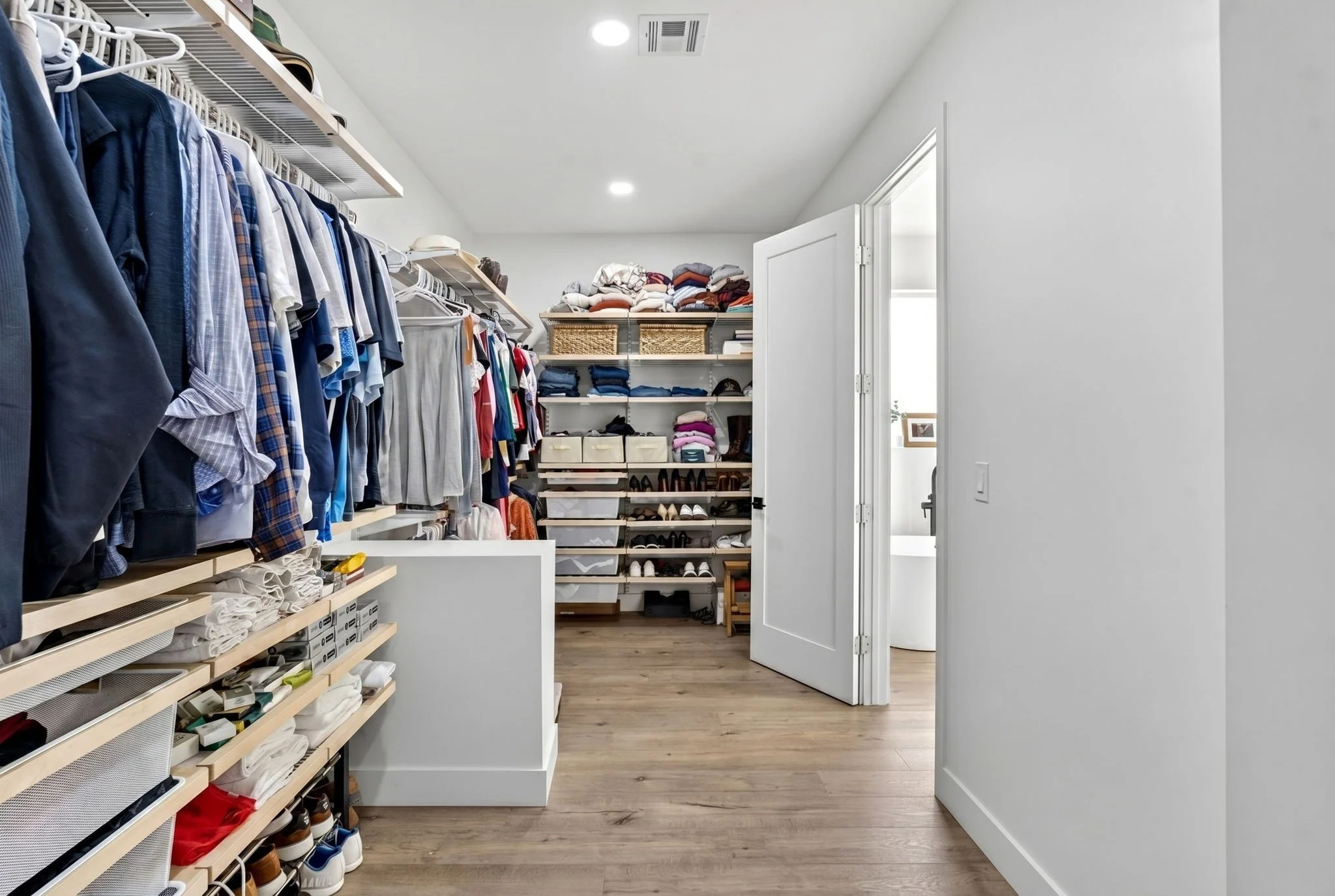 A walk-in closet with organized shelves of folded clothes, shoes, and storage bins, with a doorway leading to another room.