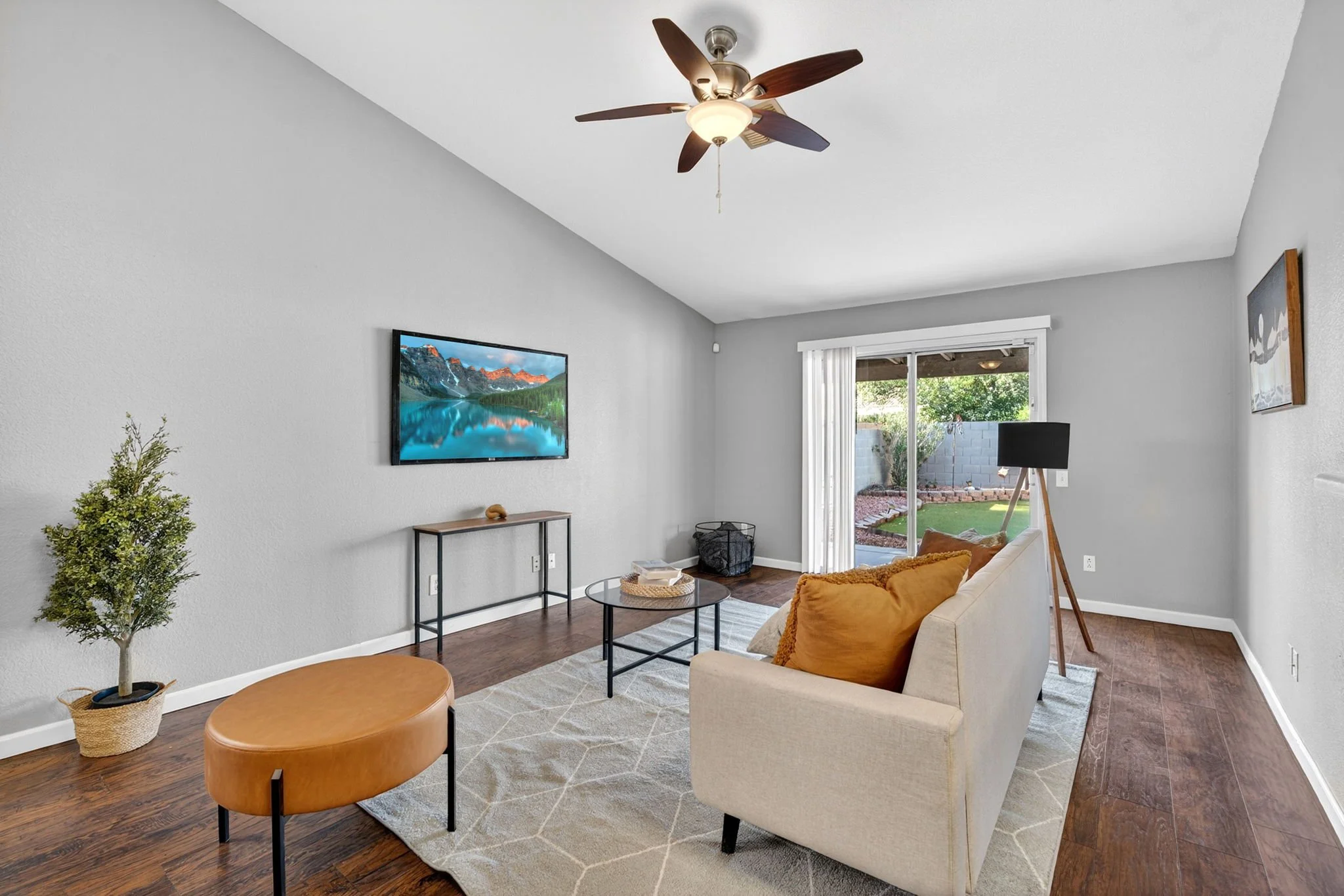Living room with beige sofa, round coffee table, potted tree, wall-mounted television, sliding door with vertical blinds, ceiling fan with light, and a window with a view of a backyard.