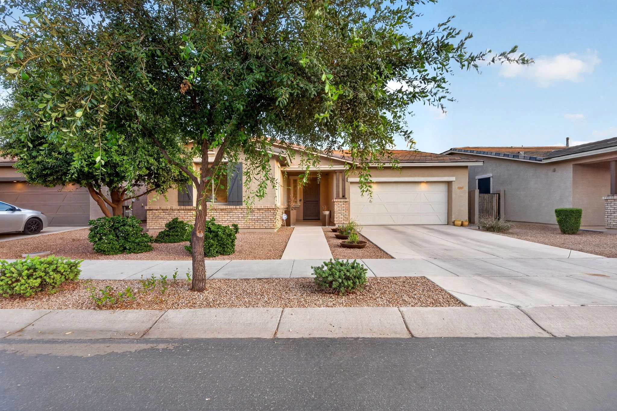 A suburban house with a tree in the front yard, a driveway with a garage, a sidewalk, and a parked car on the street.