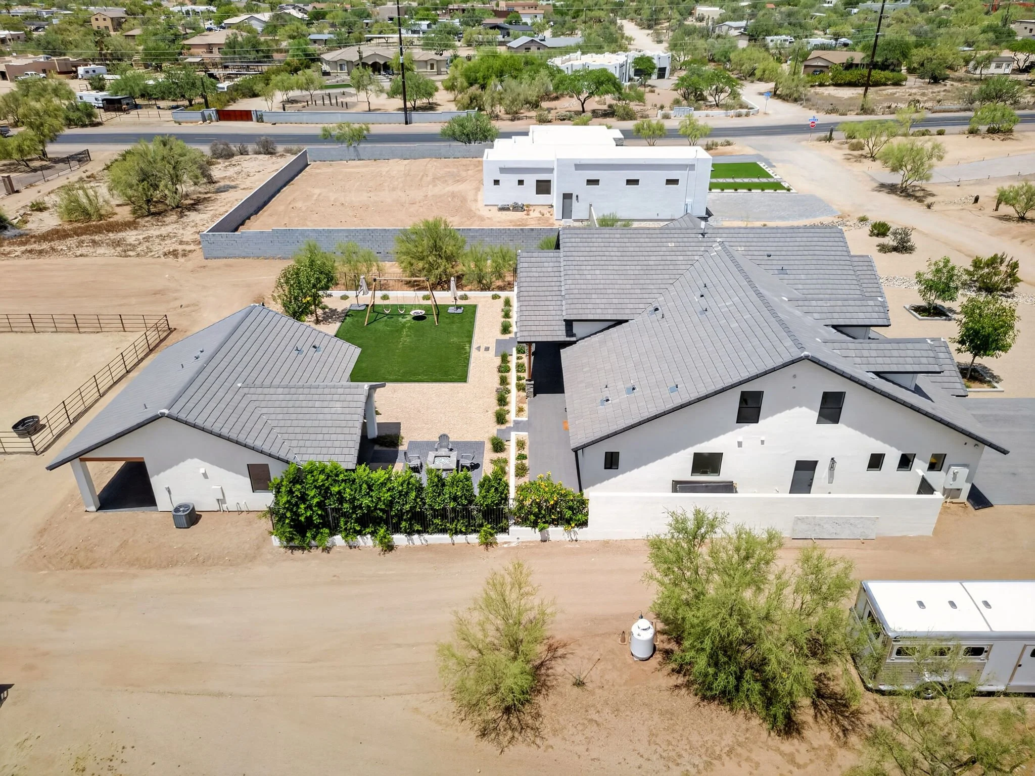 Aerial view of a residential area with newly constructed modern house, a backyard with grassy area, swings, and trees, surrounded by other homes and desert landscape.