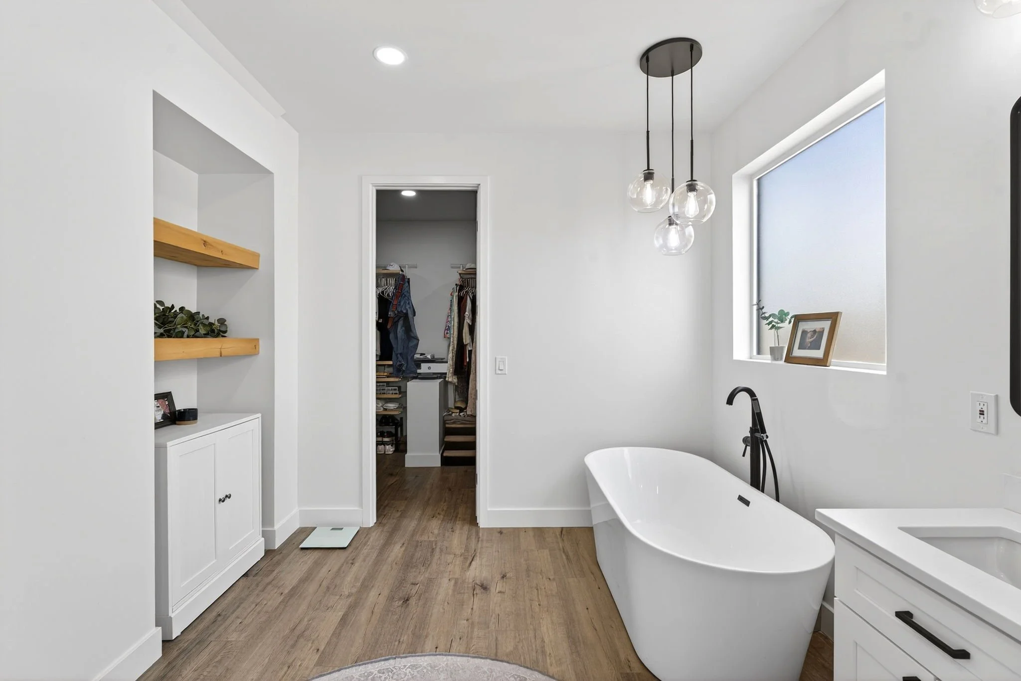 A modern bathroom with white walls, a freestanding bathtub, wooden flooring, a window with a framed photo and plant, and a walk-in closet in the background.